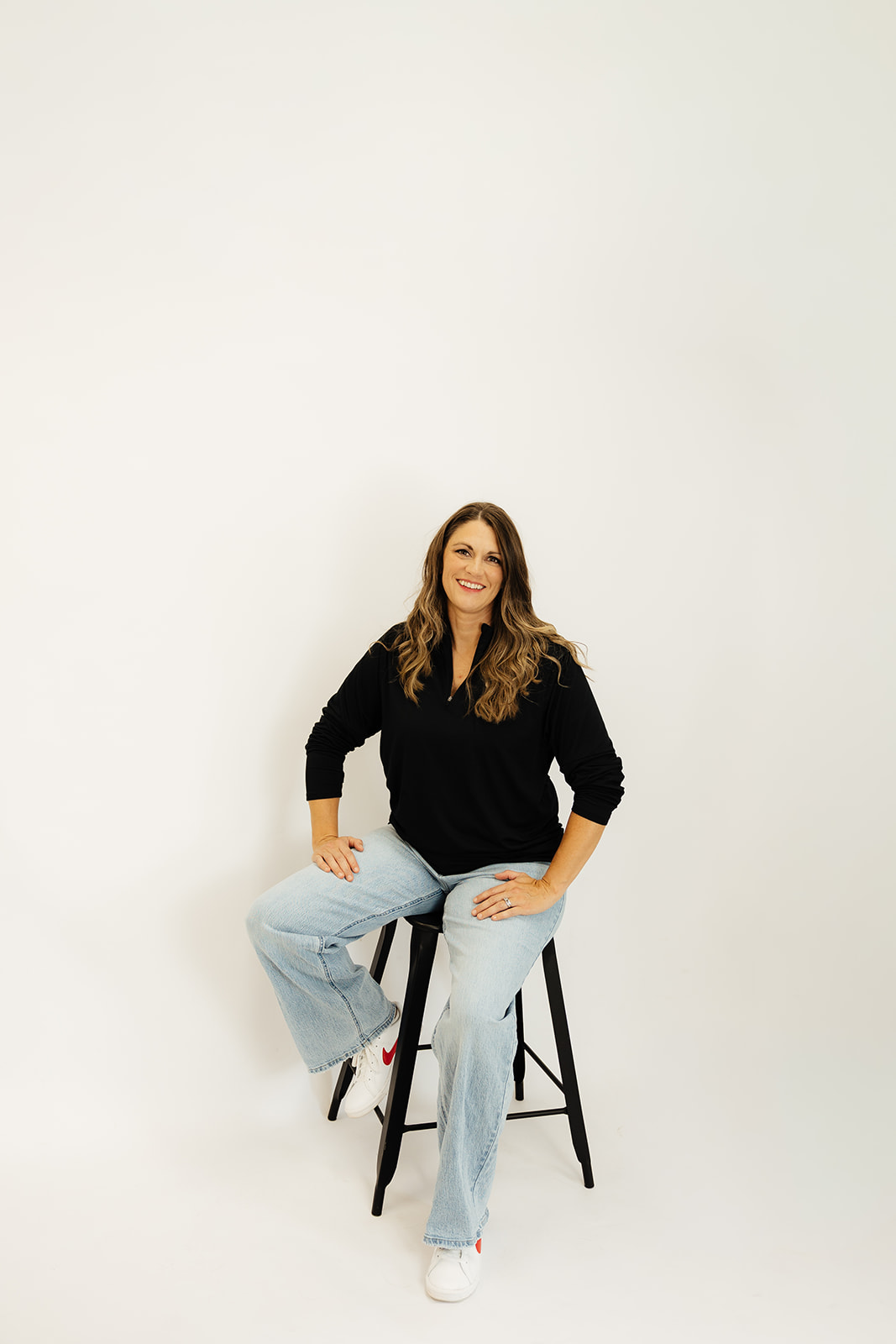 Woman with long hair sitting on a stool, smiling, wearing a black top and light blue jeans, against a plain white background.