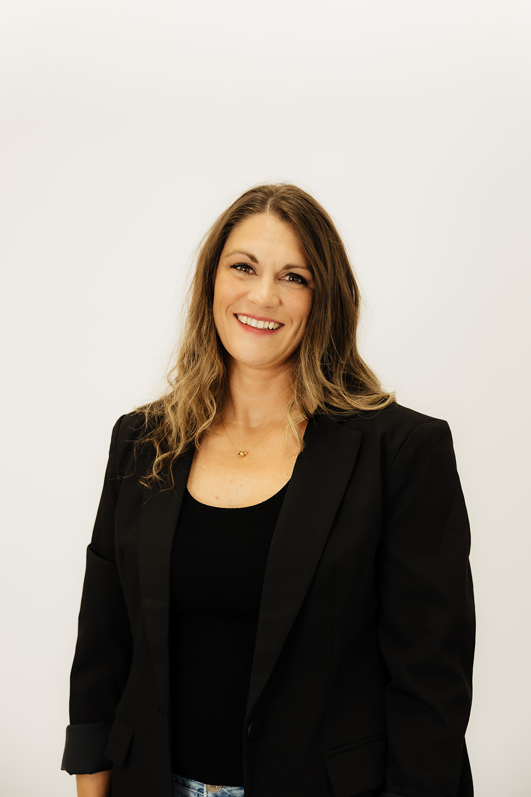 Woman with wavy hair smiling, wearing a black blazer and top, standing against a plain white background.
