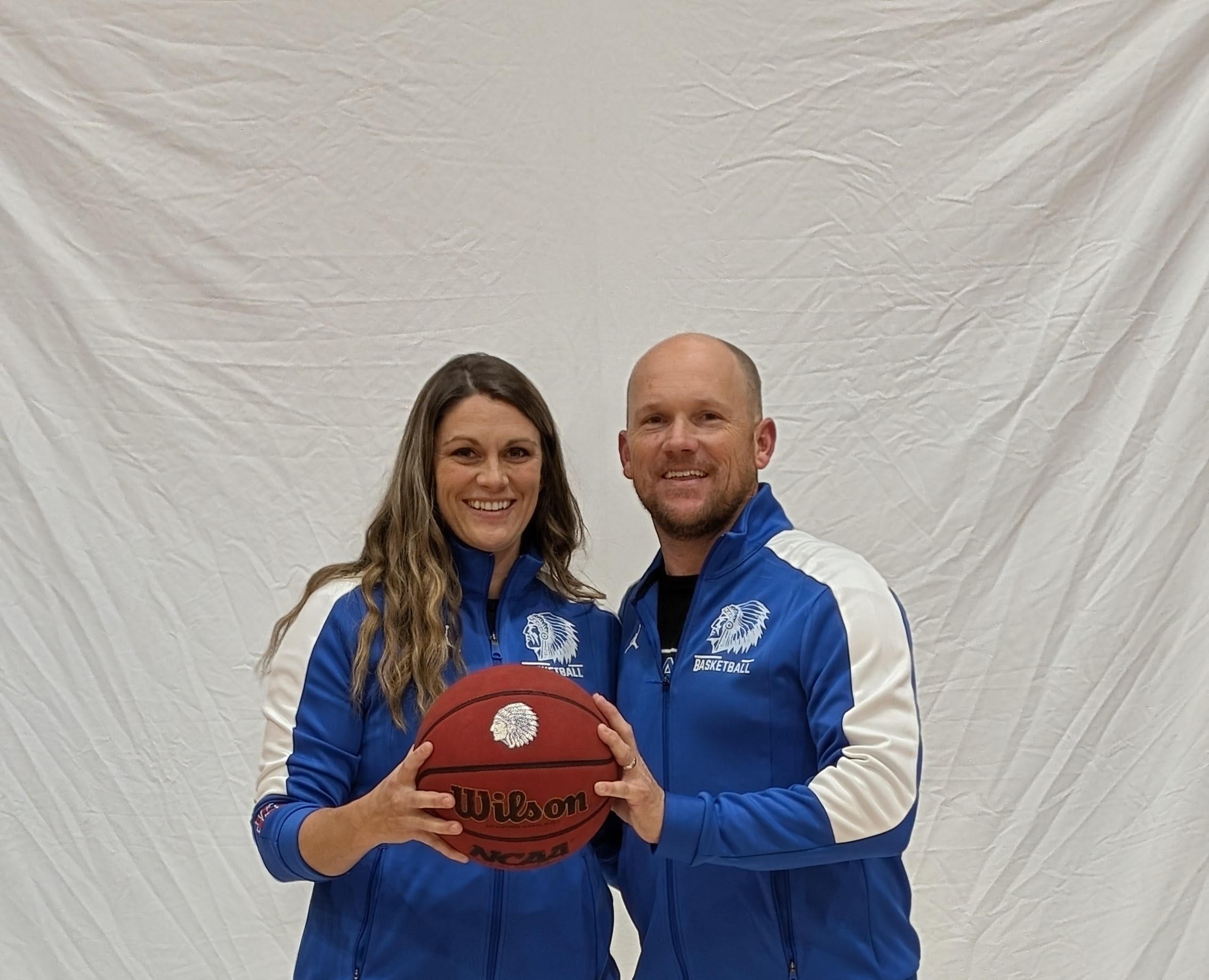 Two people in blue sports jackets holding a basketball, standing in front of a plain white backdrop.