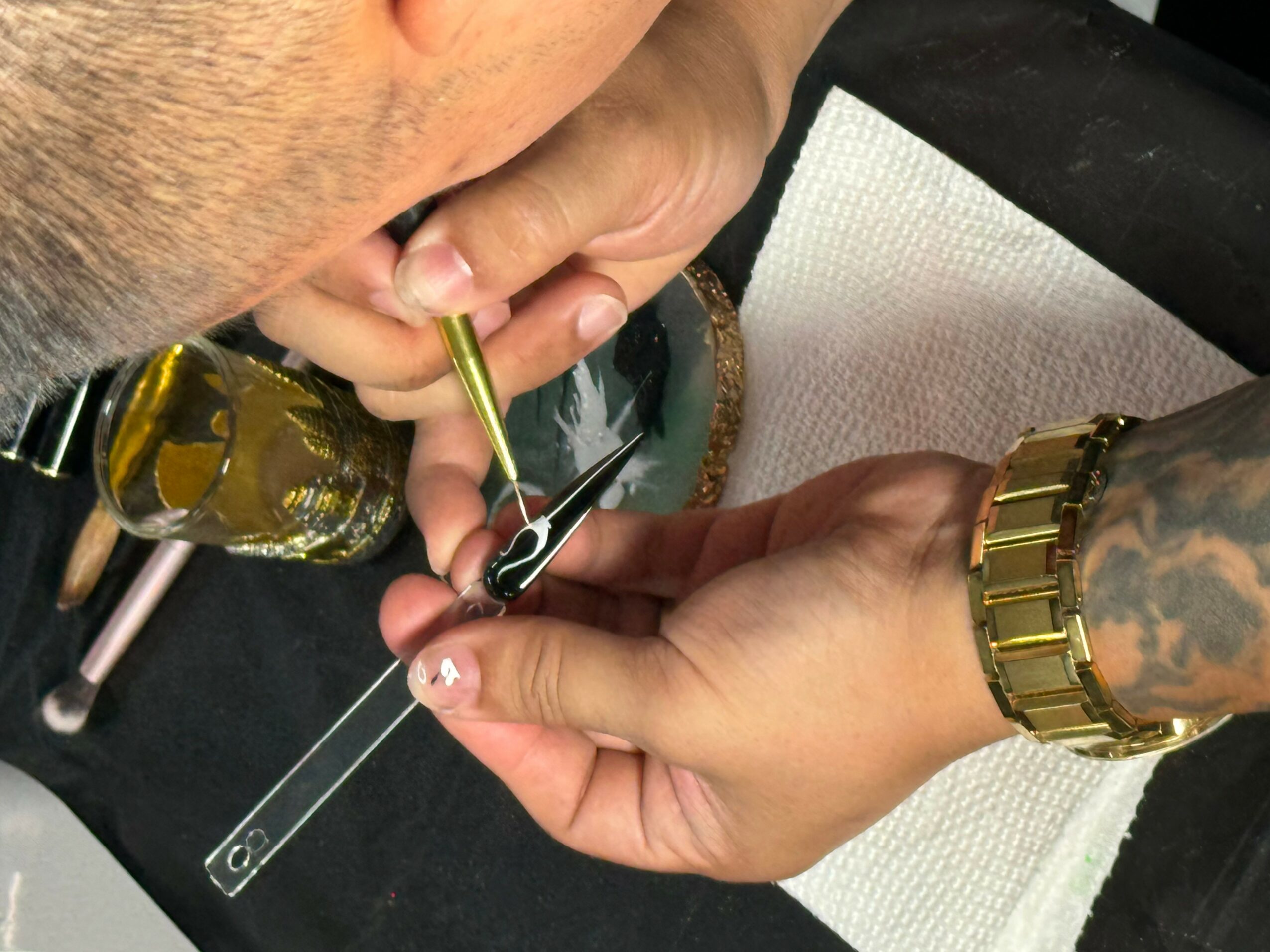 Person using a tool to work on a small object, with a gold watch and tattooed arm visible, on a black surface.
