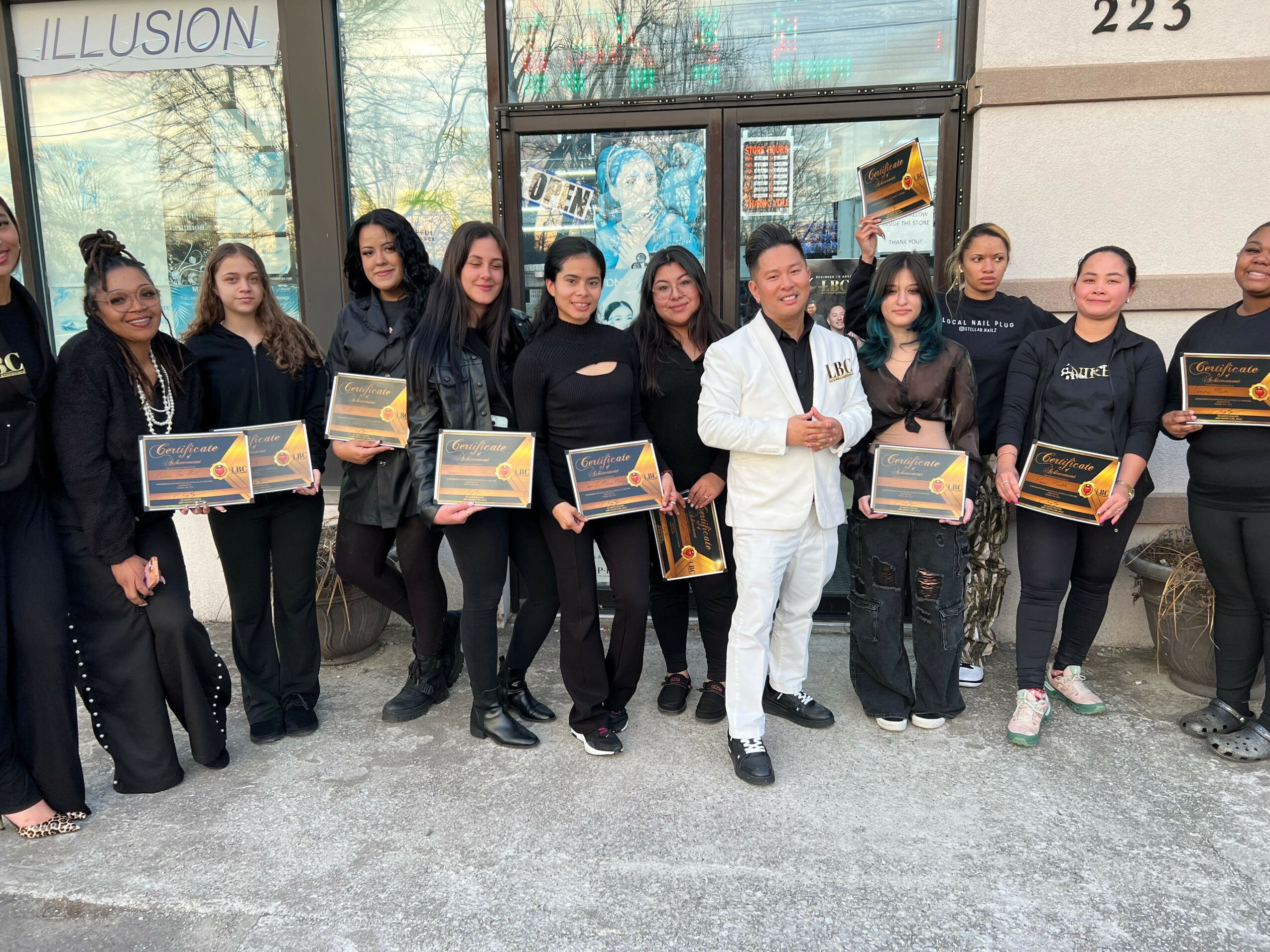 Group of people standing outside a building, holding awards or certificates, with a man in a white suit in the center.