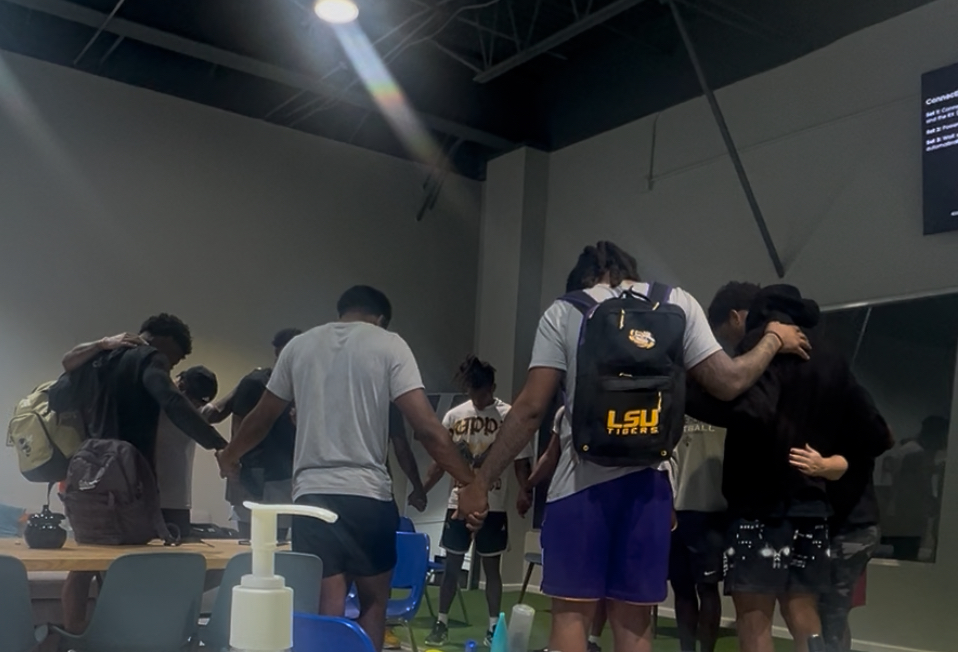 Group of people holding hands in a circle indoors, some wearing backpacks, with tables and chairs nearby.