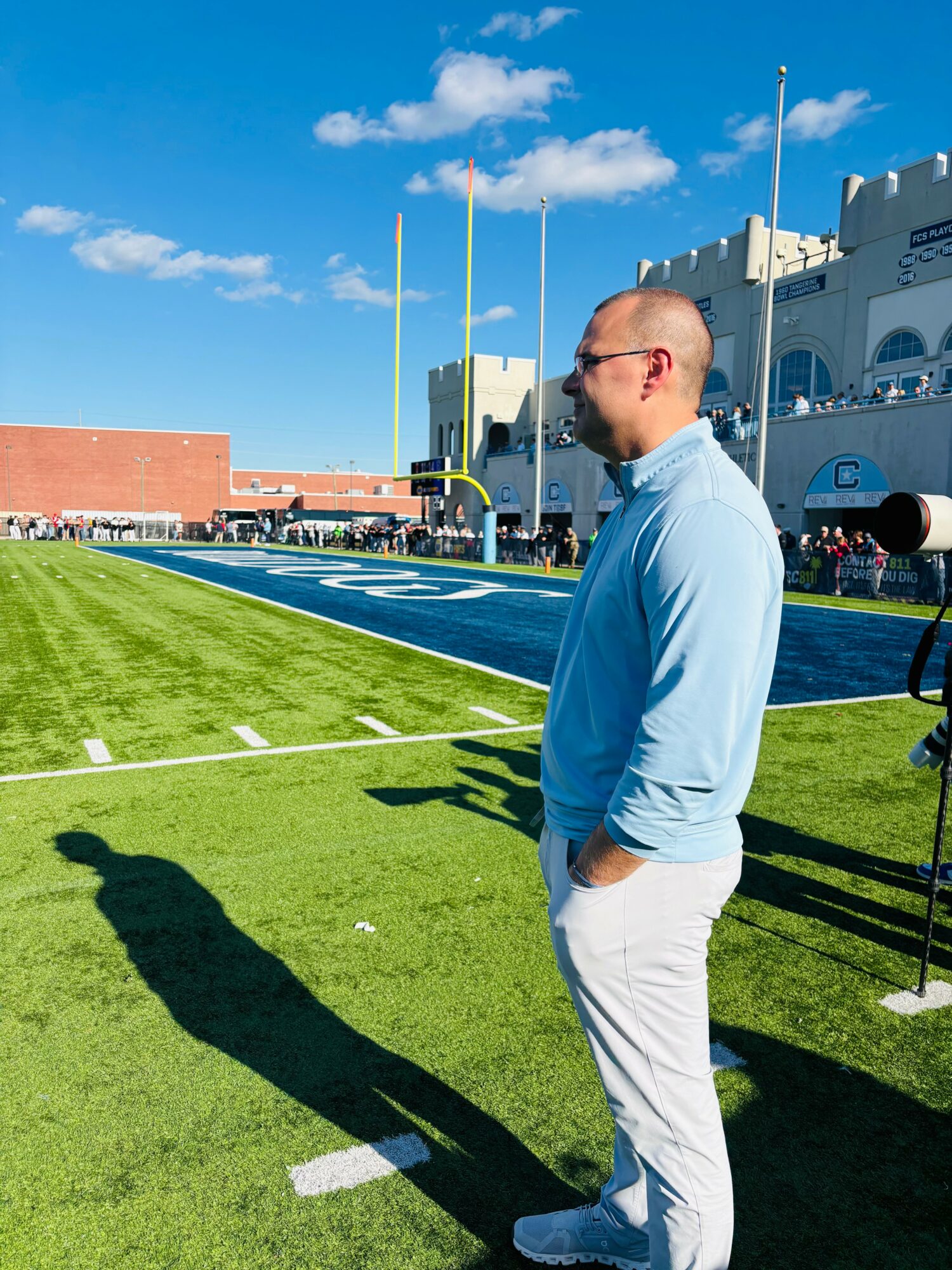 Man in light blue shirt and white pants standing on a football field with goalposts in background, sunny day.