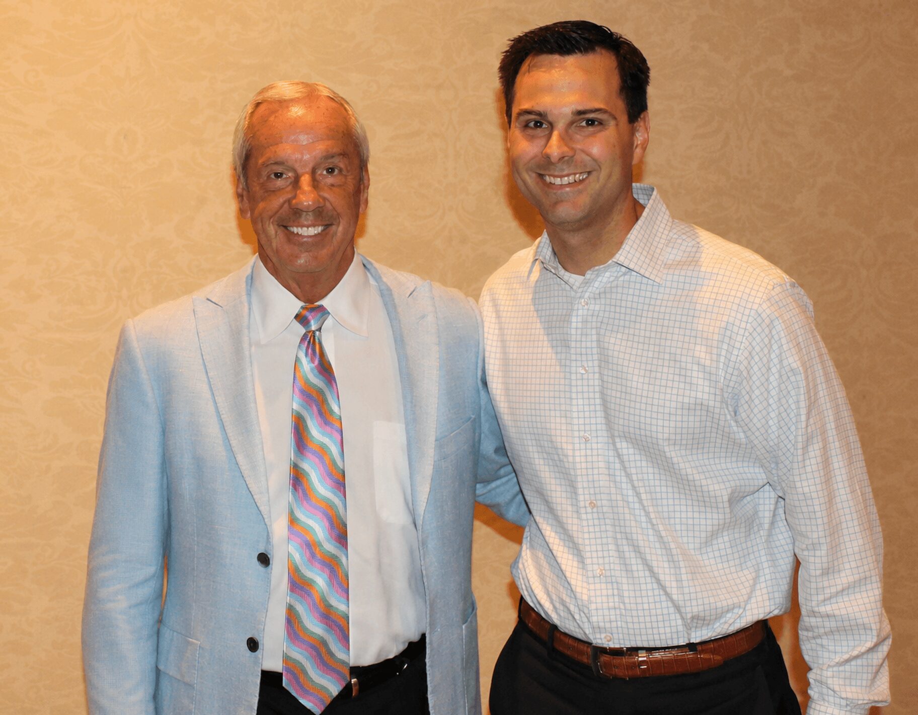 Two men standing side by side, smiling, in a beige room. One man has white hair and a colorful tie. The other has dark hair and a light shirt.