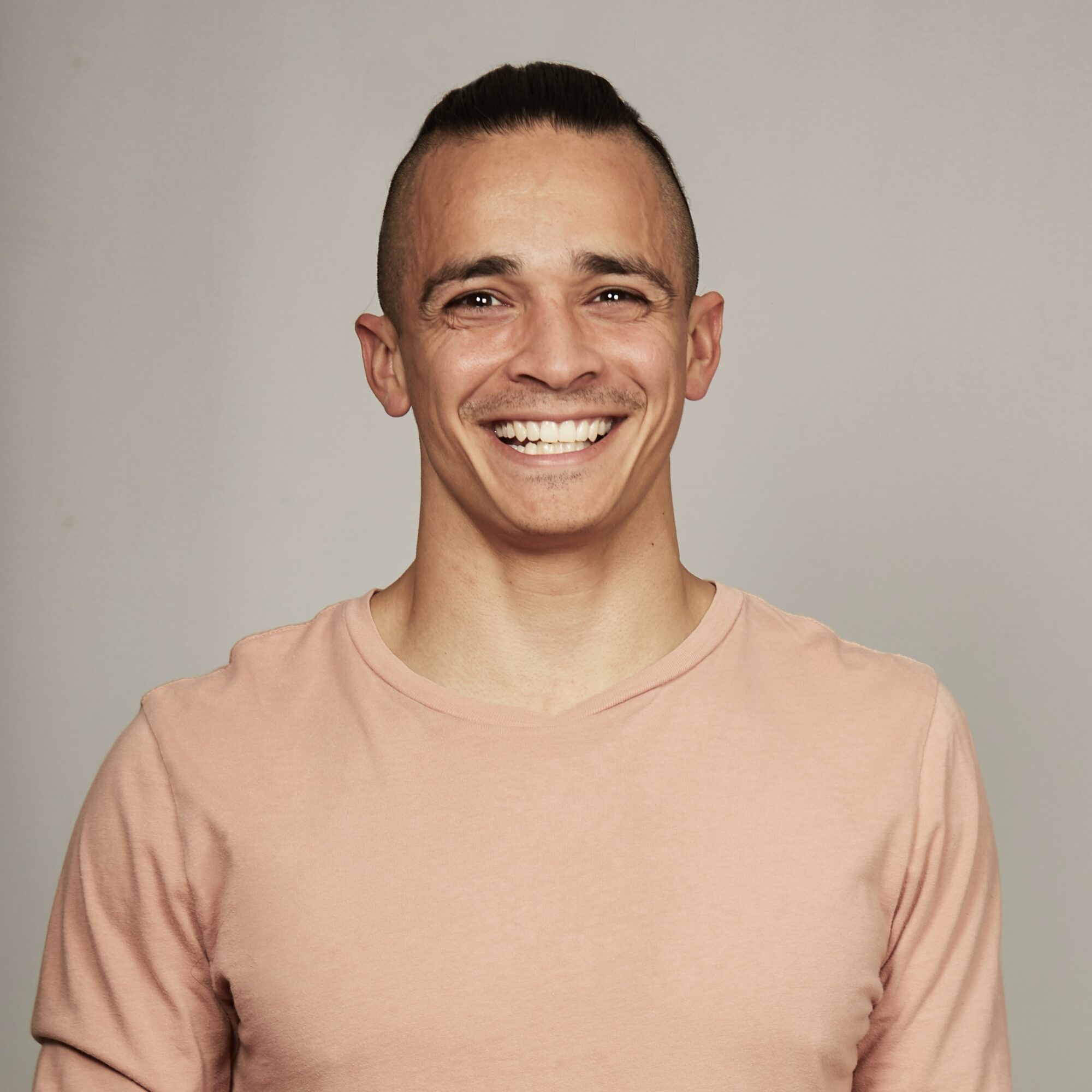 Smiling man with short dark hair and a light pink shirt, facing forward, neutral background.