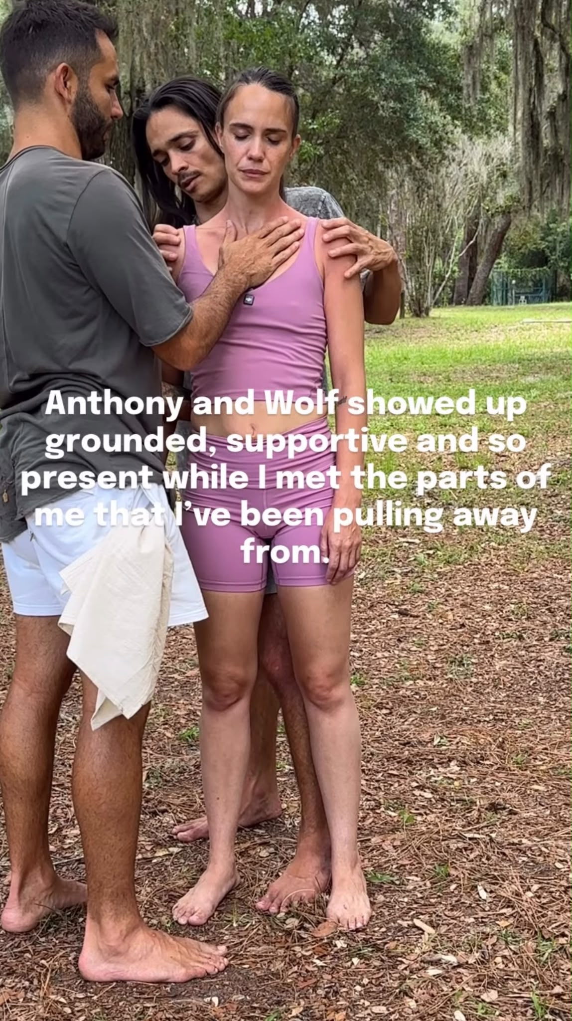 Three people standing outdoors, supporting a woman in a pink top and shorts, with trees in the background.