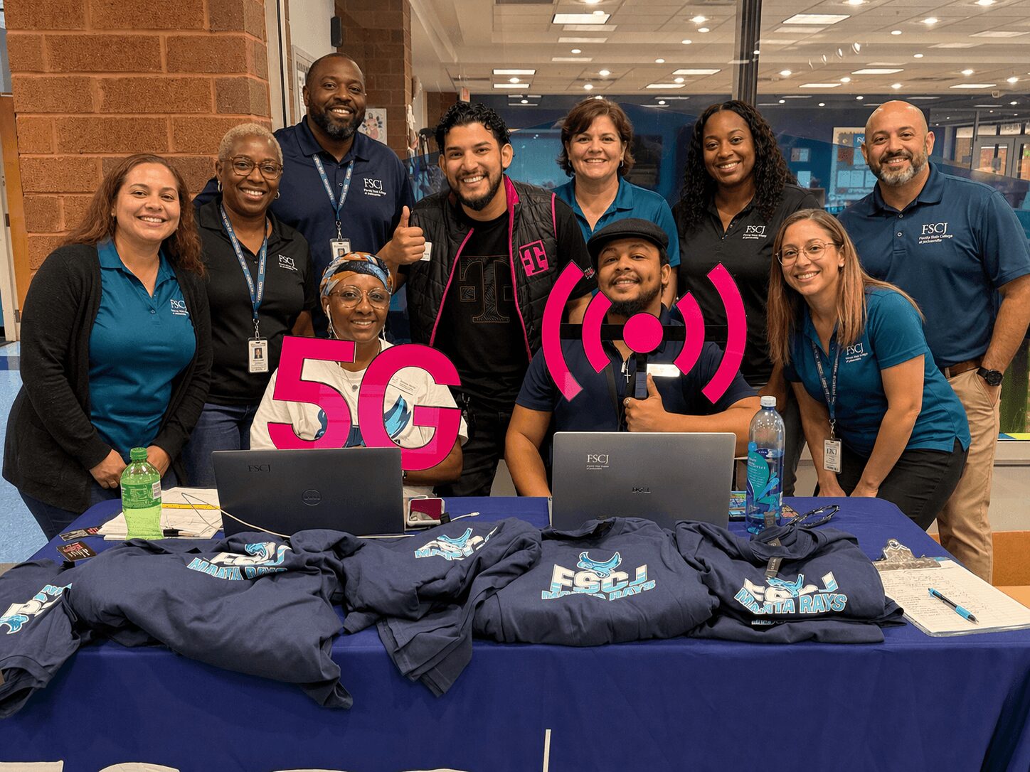 Group of ten people smiling behind a table with laptops and 5G signs, indoors in a modern building.