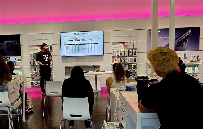 Person presenting in front of a screen with seated audience in a store or showroom, pink lighting overhead.