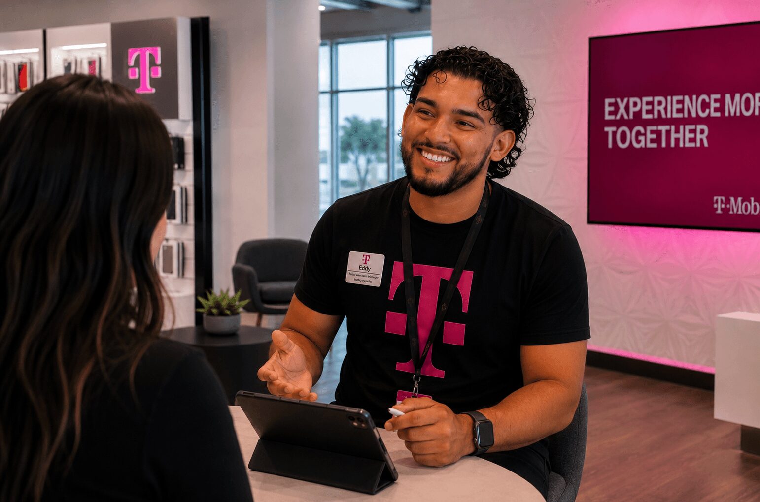 A man with curly hair and a beard smiling while talking to a woman at a T-Mobile store counter.