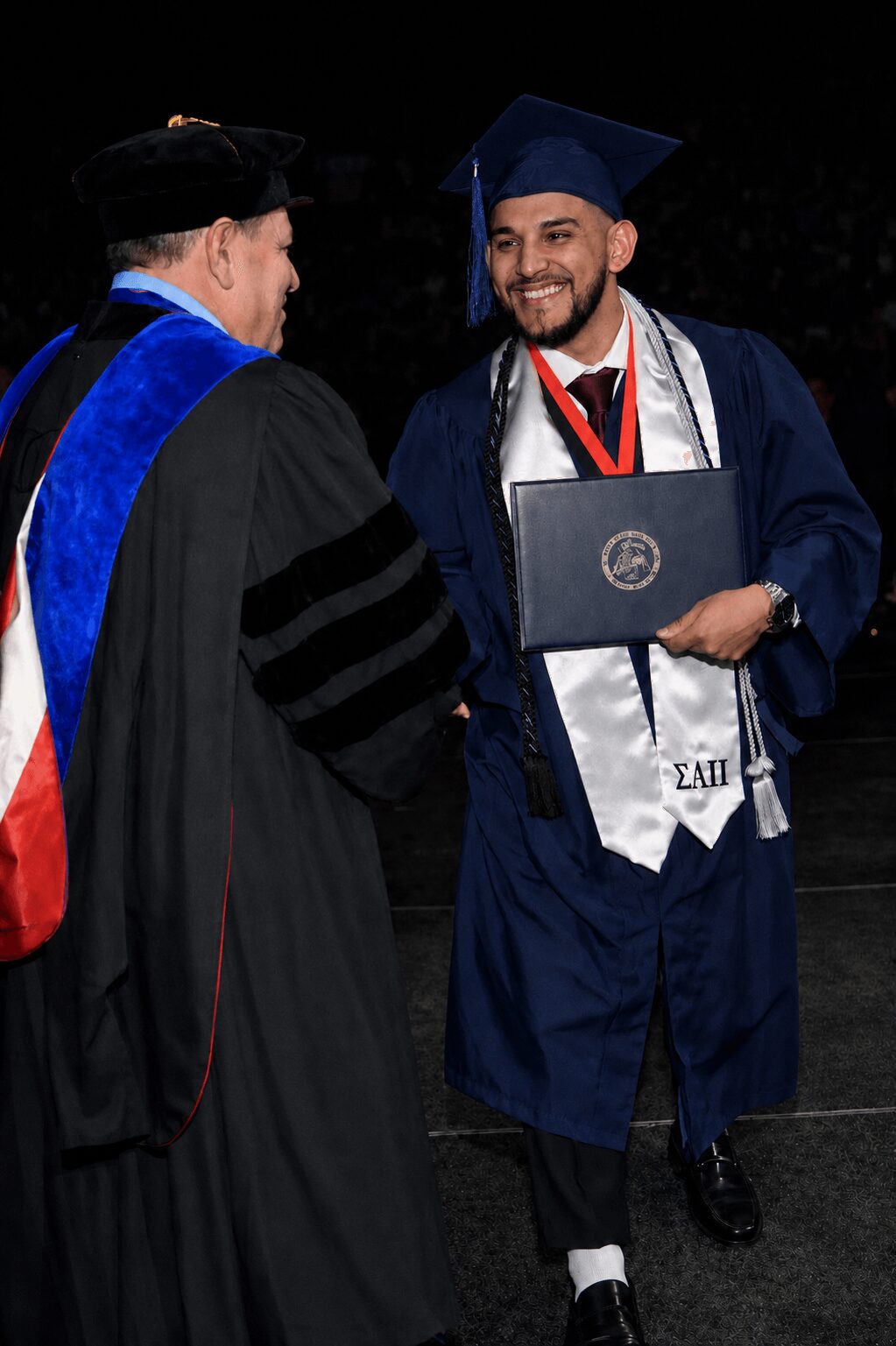 Graduate in blue cap and gown receiving diploma from faculty member in academic regalia, smiling, dark background.