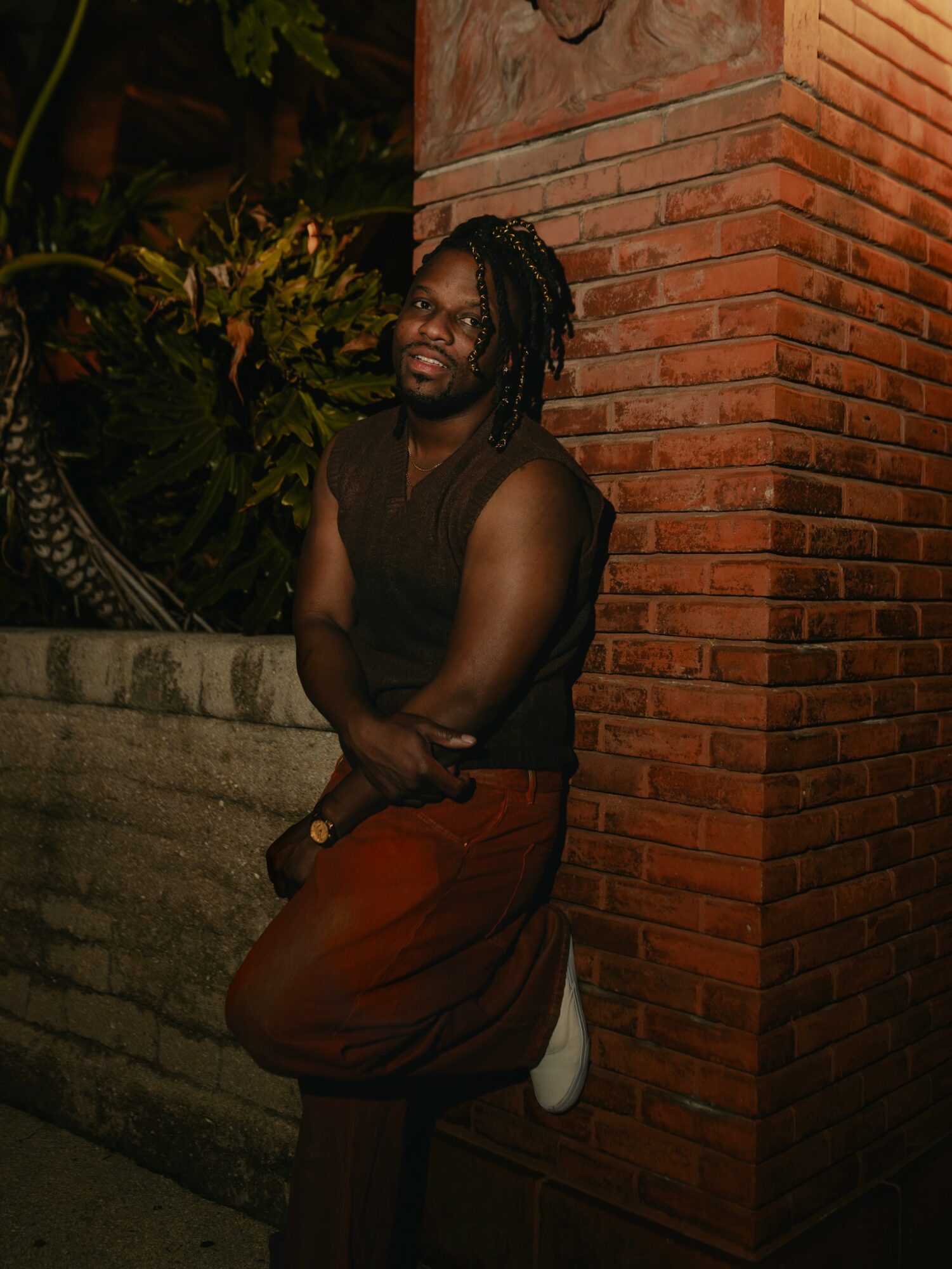 Person with dreadlocks leaning against a brick wall outdoors at night, smiling, wearing a sleeveless top and brown pants.