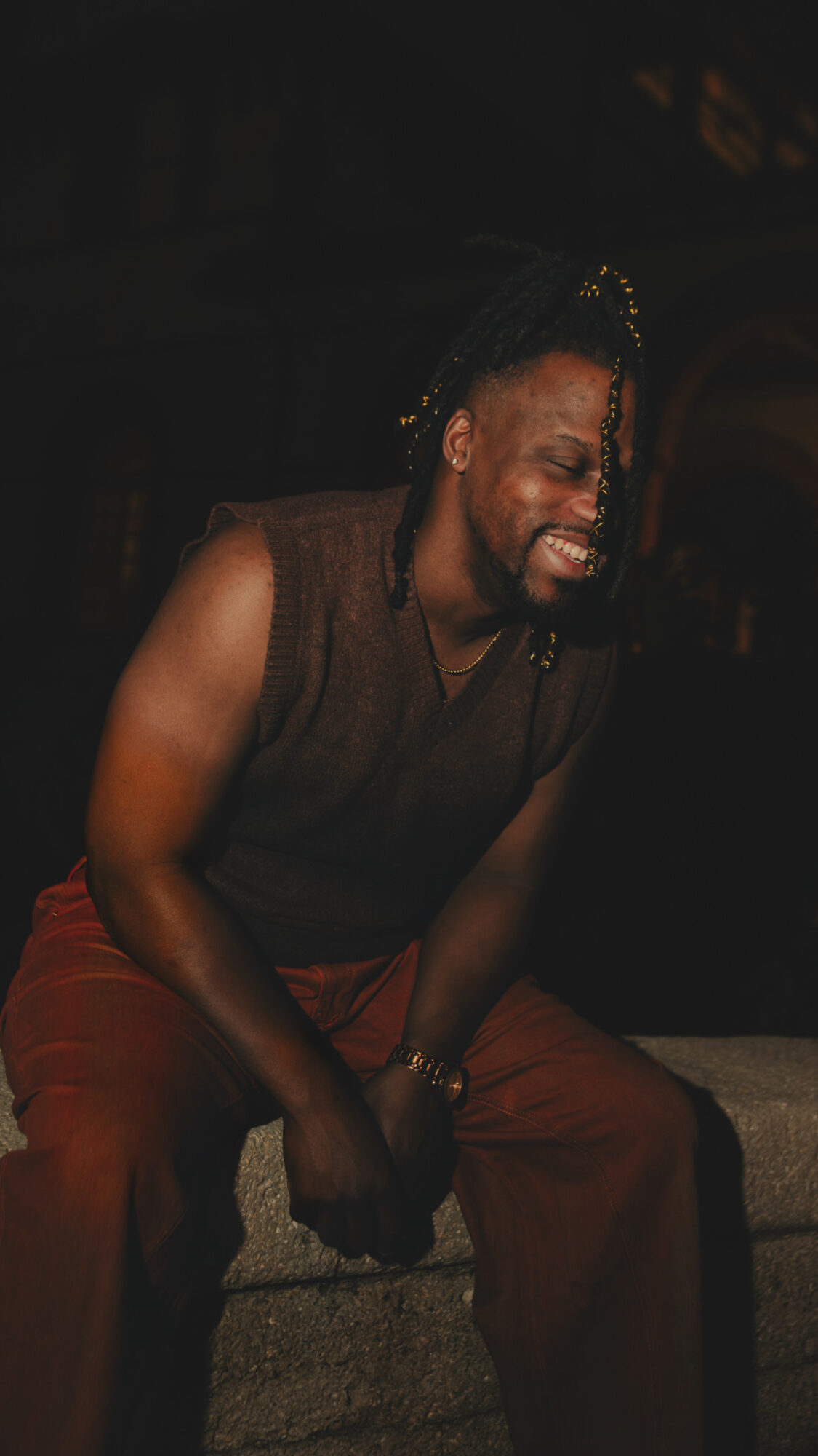 Person laughing with eyes closed, wearing a sleeveless top and red pants, sitting on a bench in a dark setting.
