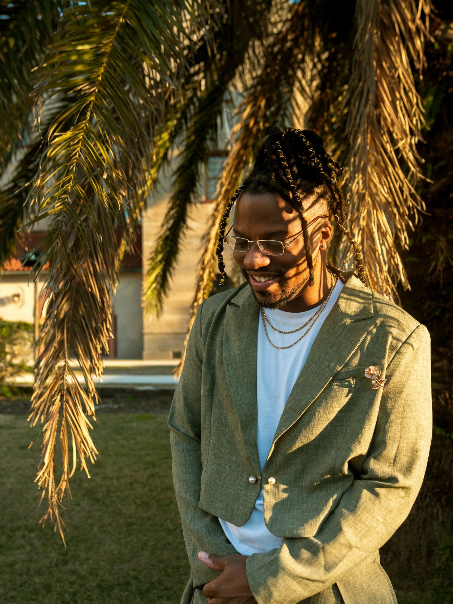 Man with glasses and braided hair smiling outdoors near palm tree, wearing a green blazer and white shirt.