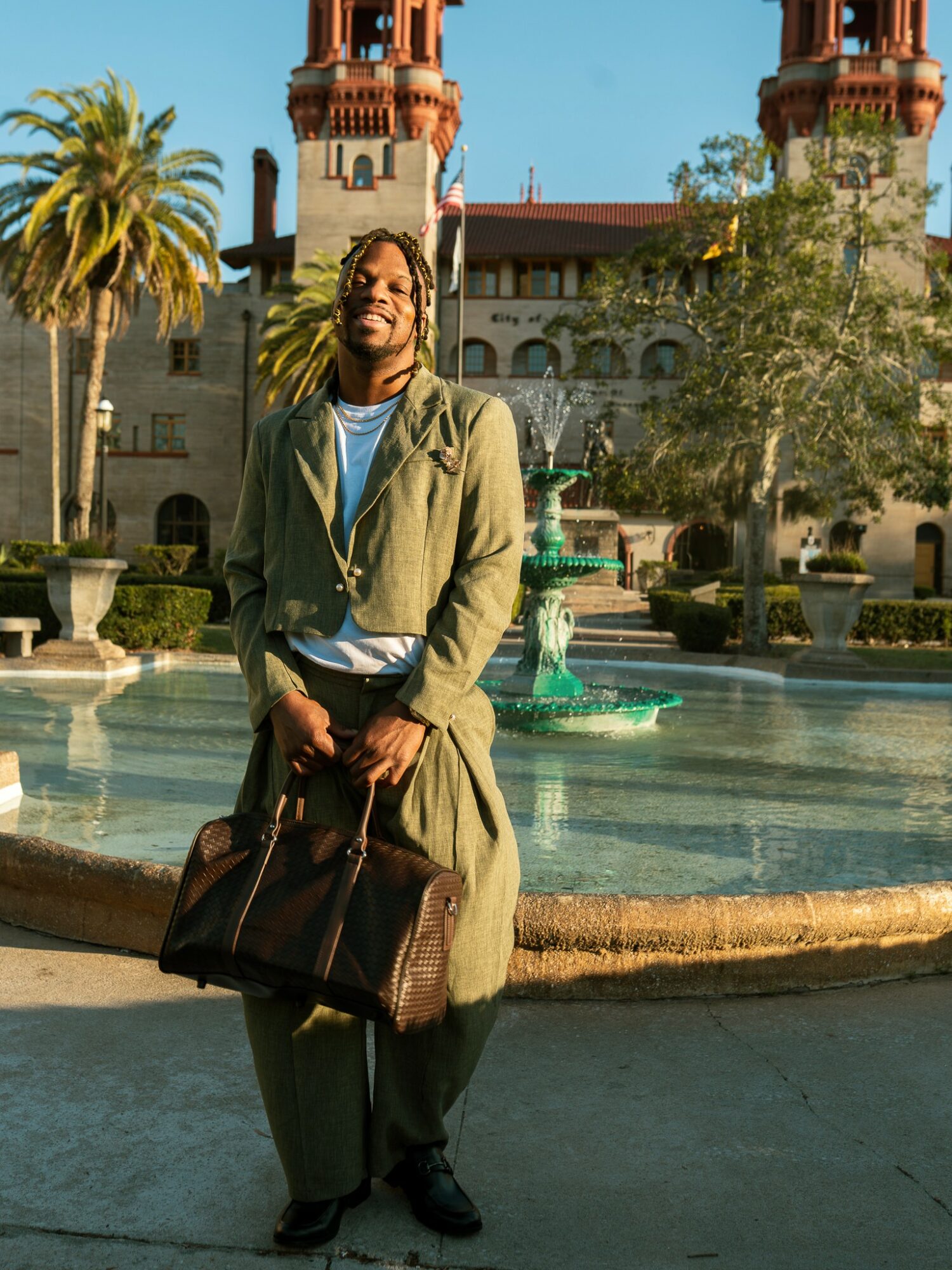 Person standing in front of a fountain with a historic building and palm trees in the background.