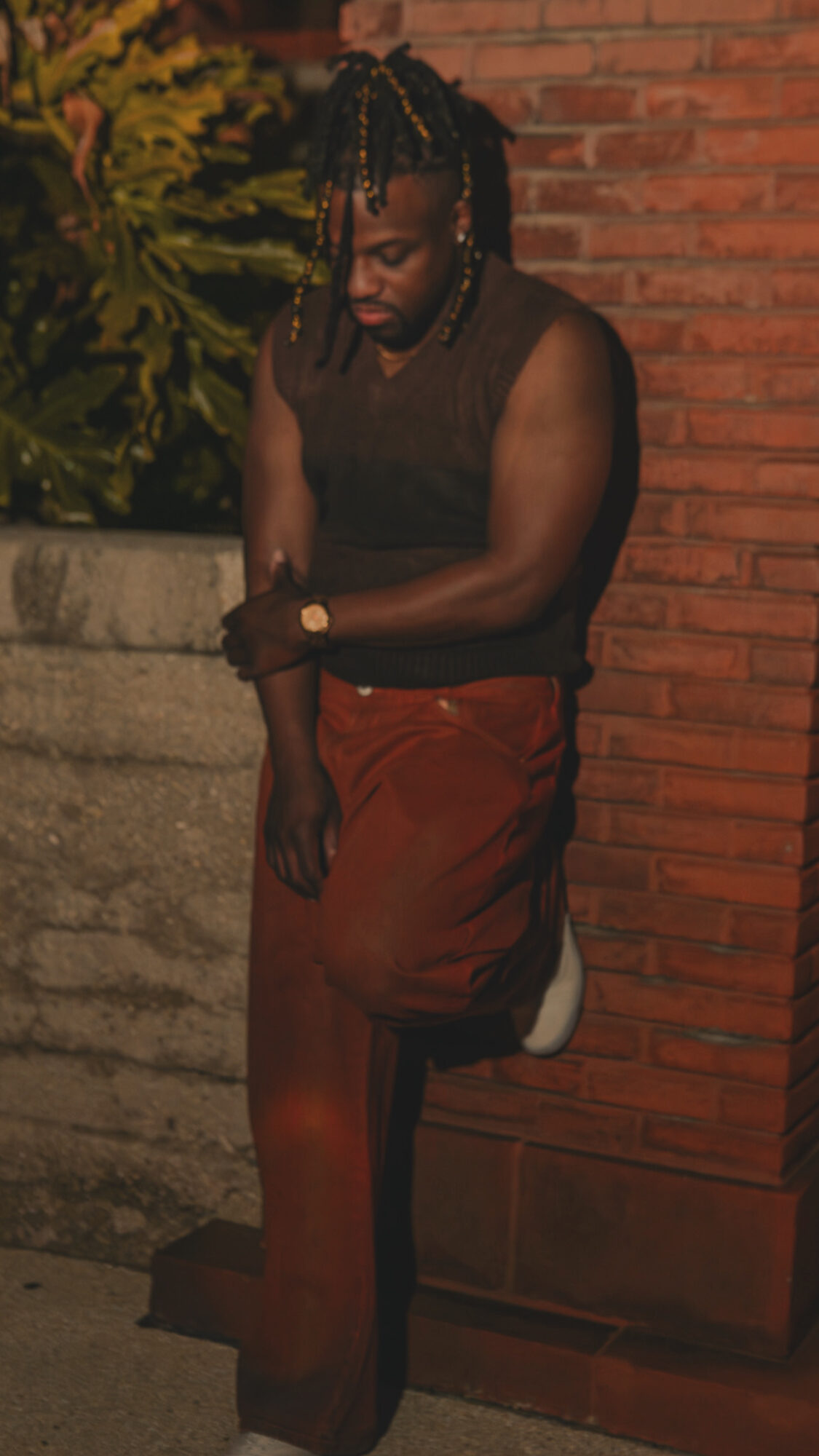 Person with braided hair leaning against a brick wall, wearing a sleeveless top and red pants, outdoors at night.