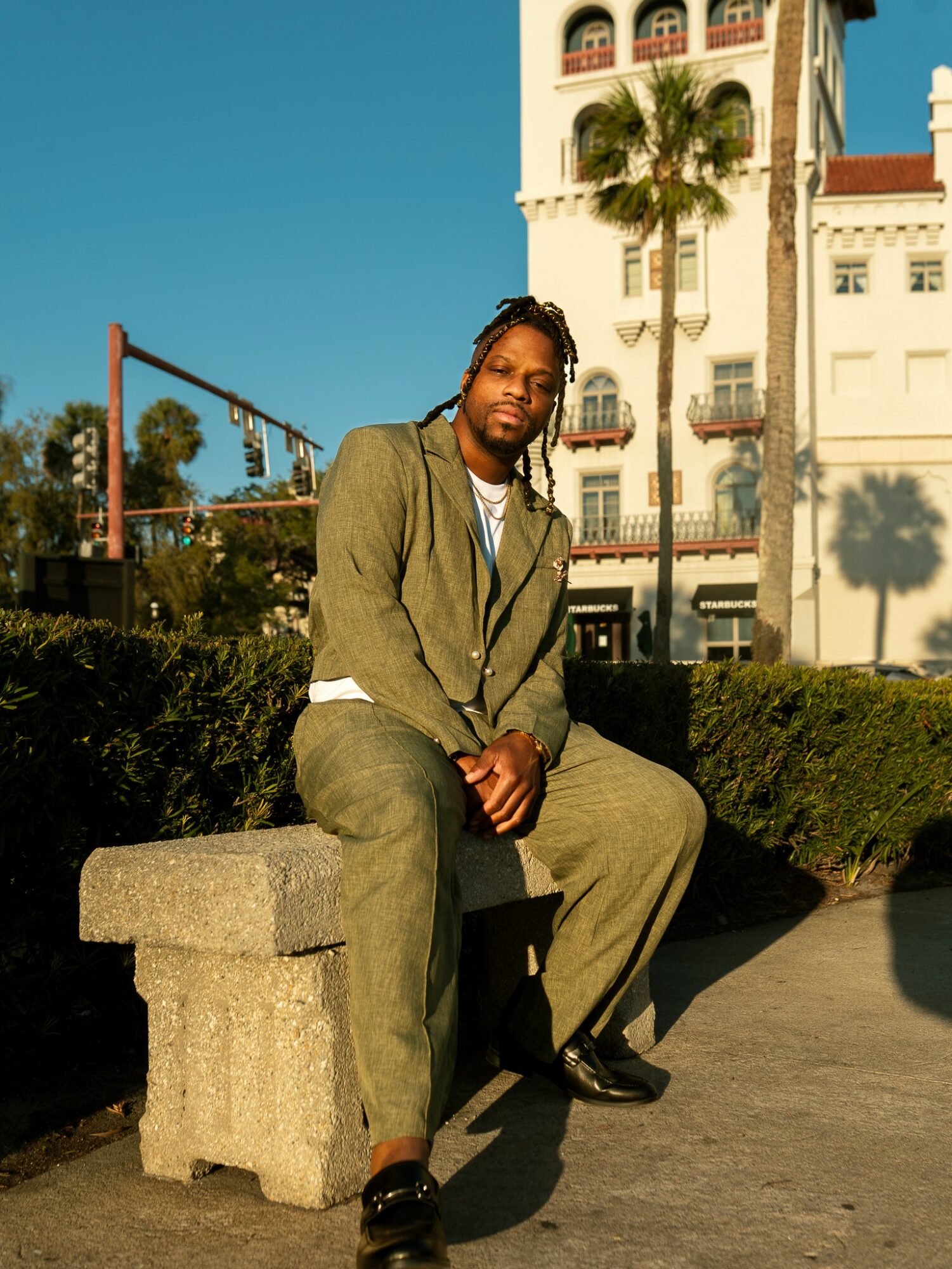 Man sitting on a concrete bench outdoors with a white building, palm trees, and a clear blue sky in the background.