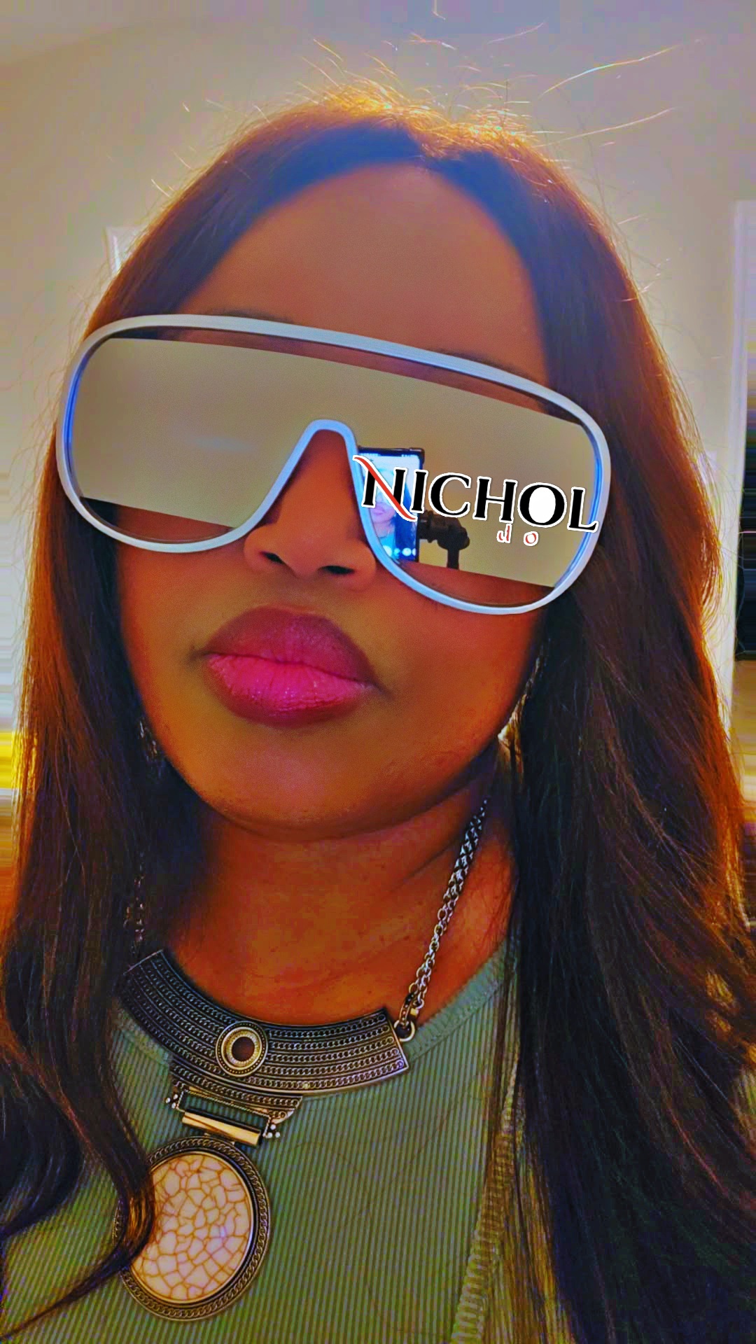 Woman wearing large sunglasses with reflective lenses, lips pursed, outdoors with sunlight, wearing a necklace and green top.