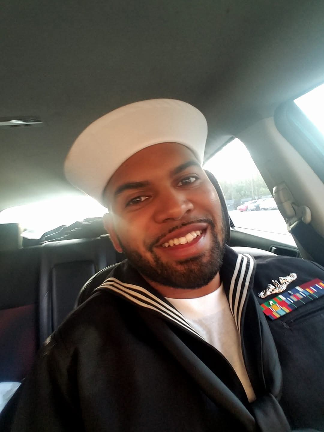 Smiling man in a navy uniform with medals, wearing a white cap, inside a vehicle.