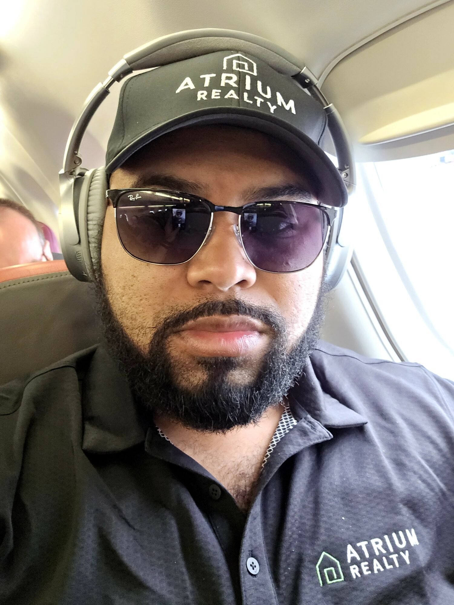 Man wearing sunglasses, a cap with 'Atrium Realty' logo, and headphones, sitting on an airplane.