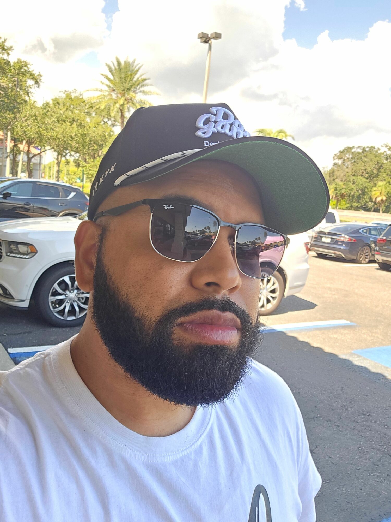 Man with beard wearing sunglasses and a cap, outdoors in parking lot, sunny weather.
