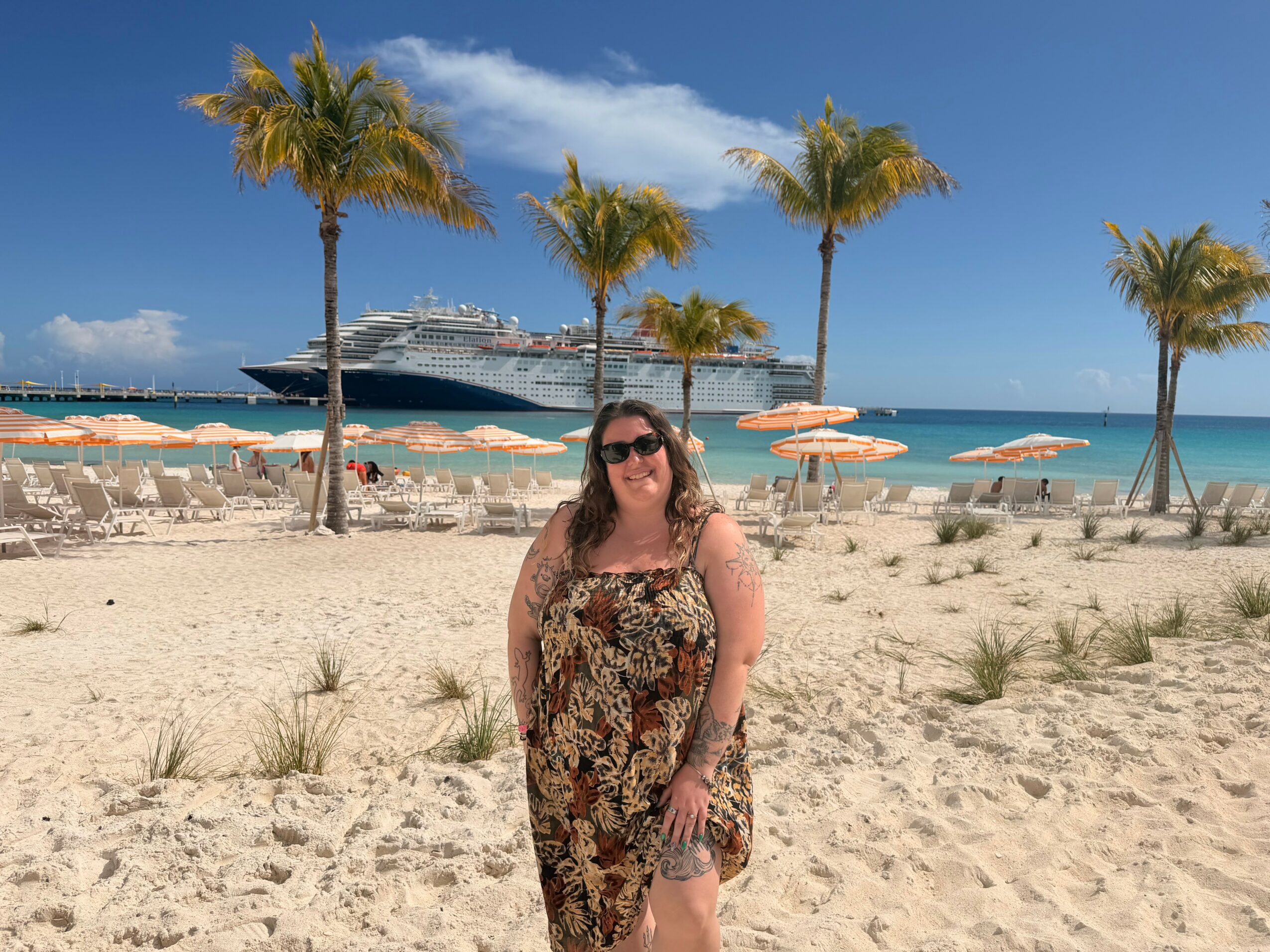 Woman in a floral dress and sunglasses standing on sandy beach with palm trees, cruise ship, and umbrellas in background.