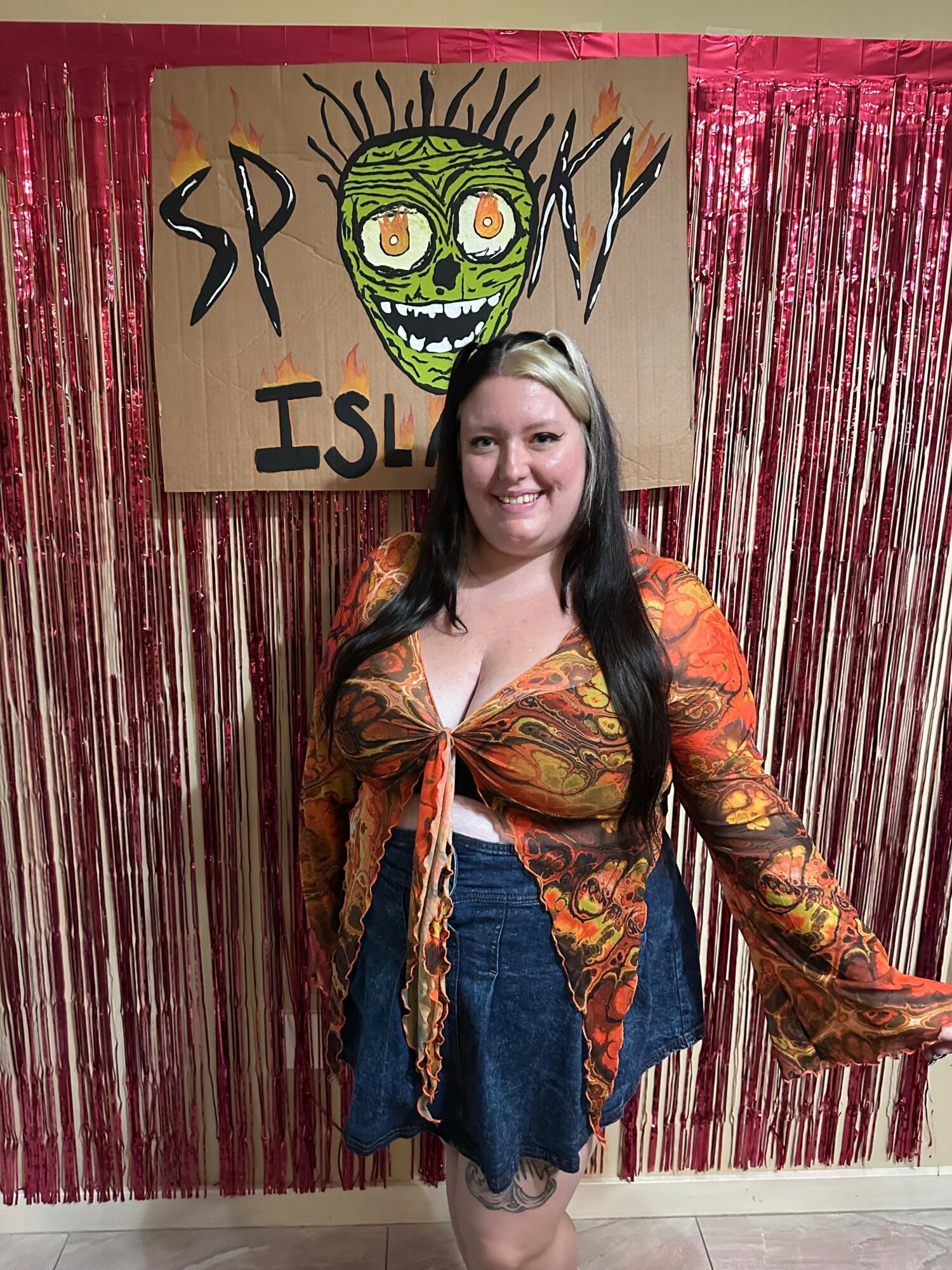 Woman standing next to a colorful sign with a green cartoon face and text, in front of a pink curtain backdrop.