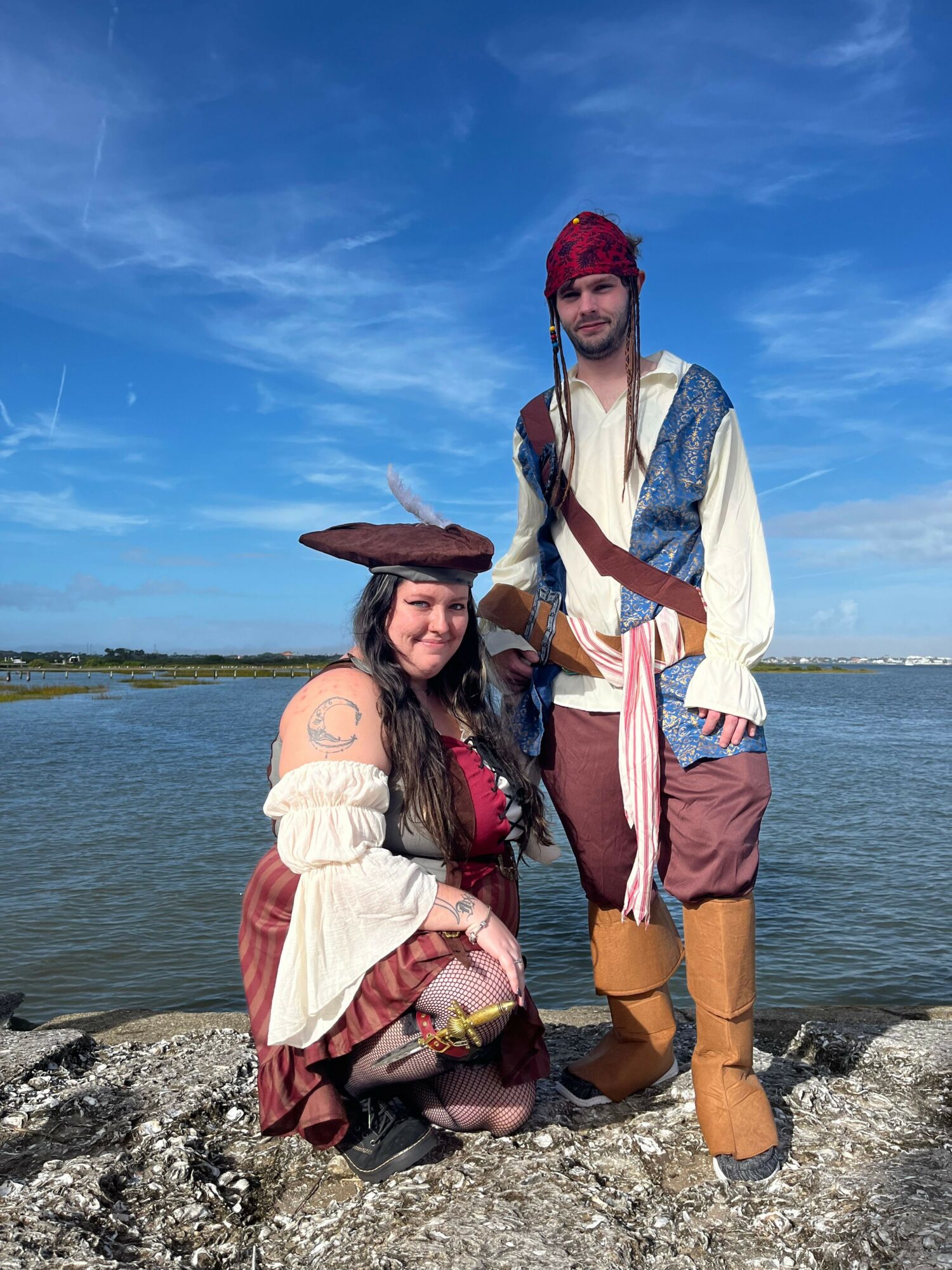Two women in historical costumes by water, one standing and one sitting on rocks, with a blue sky background.