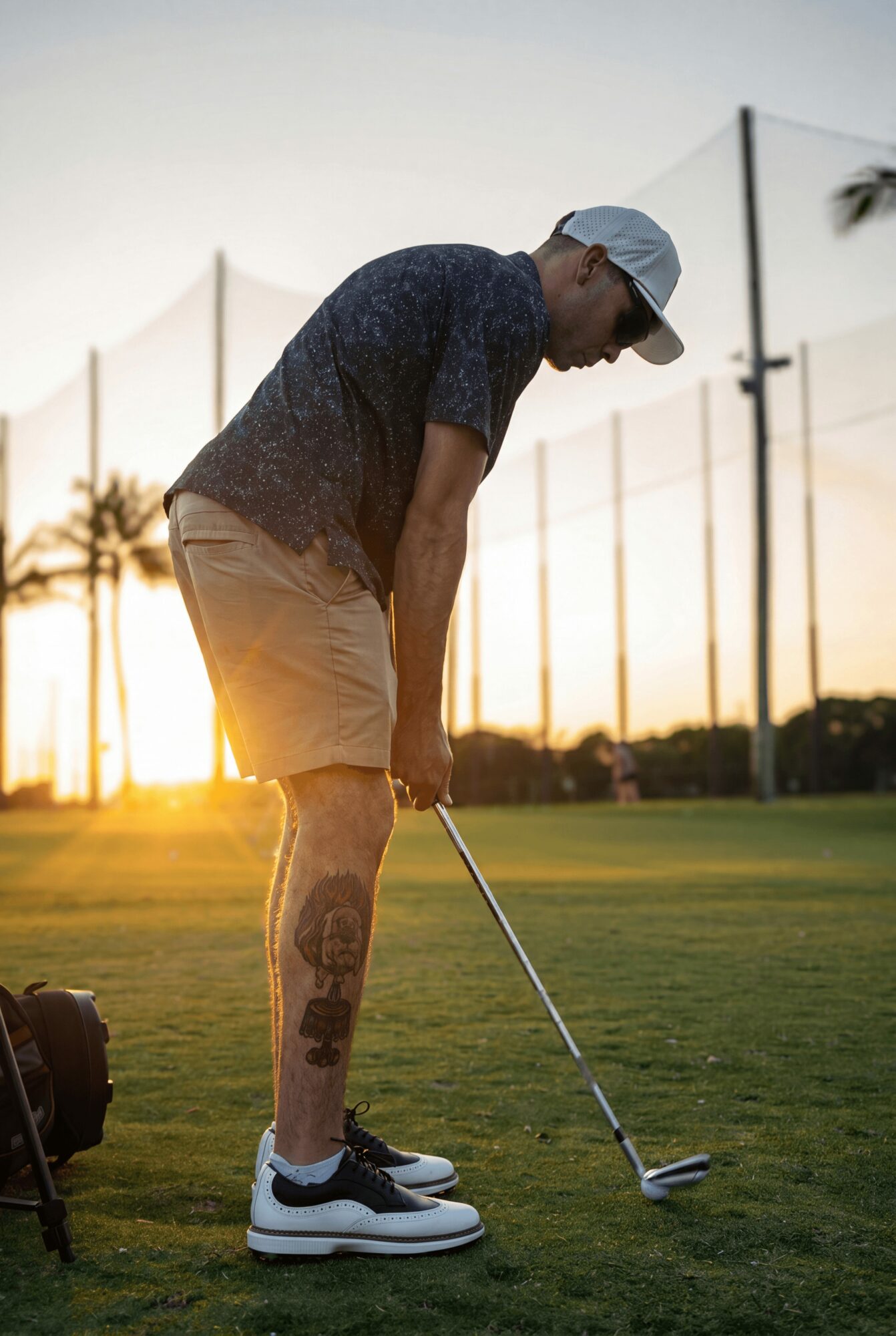 Person preparing to hit a golf ball on a field during sunset, wearing a cap, shorts, and sneakers.