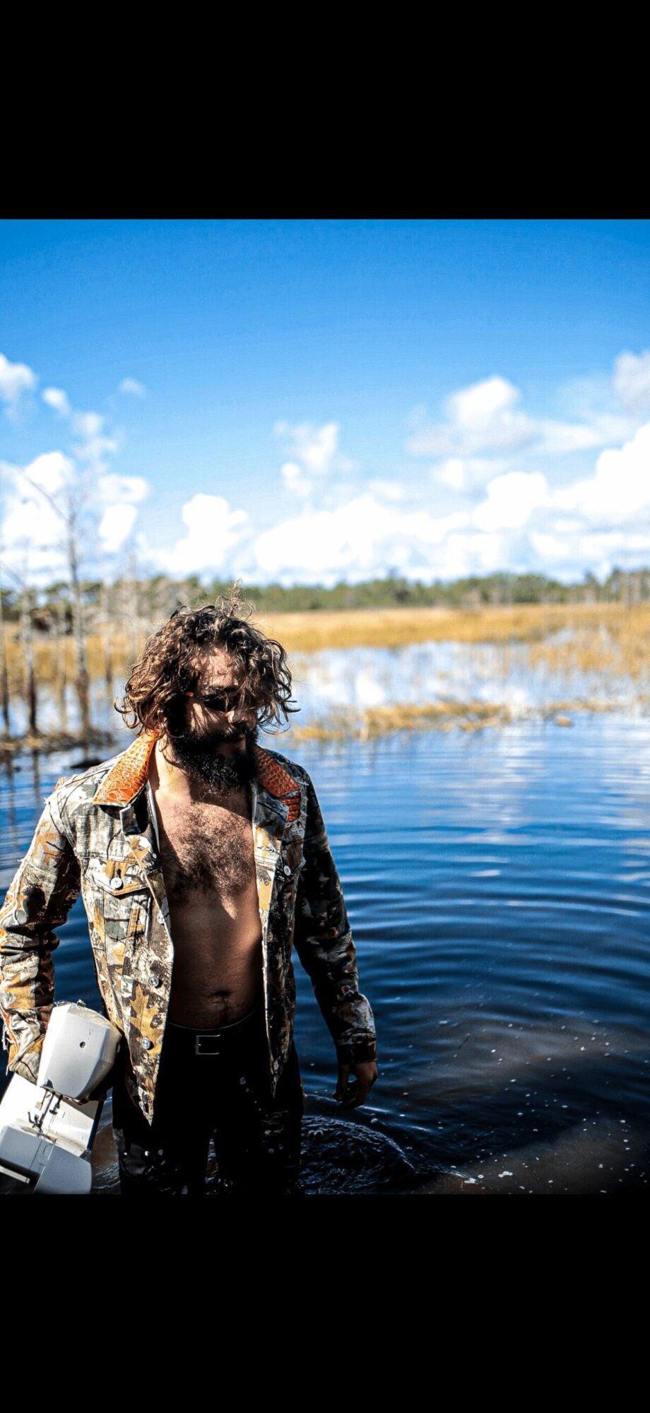 Person with curly hair and camouflage jacket standing in water outdoors under blue sky with clouds.