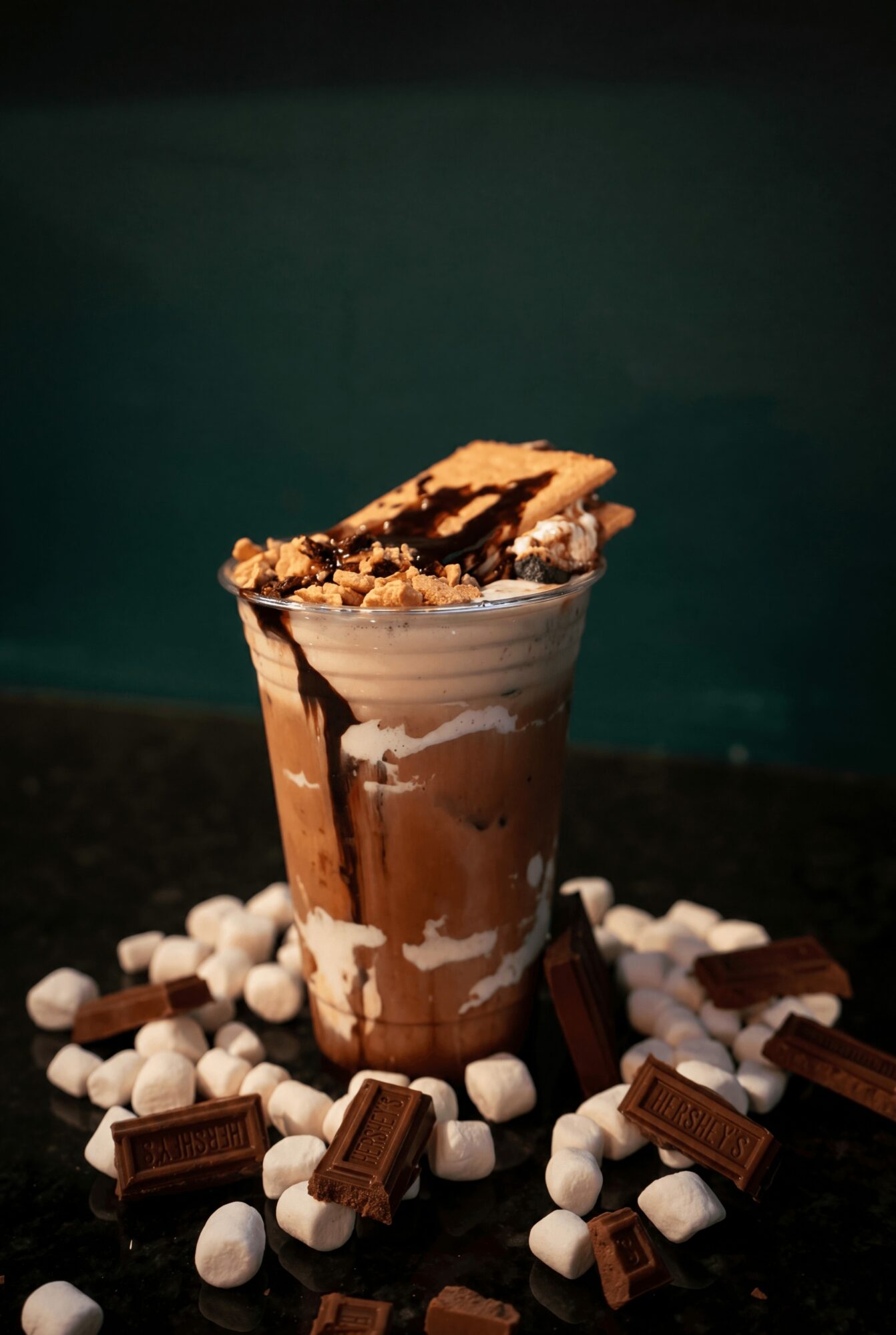 Chocolate milkshake topped with cookie pieces, surrounded by marshmallows and chocolate bars on a dark surface.