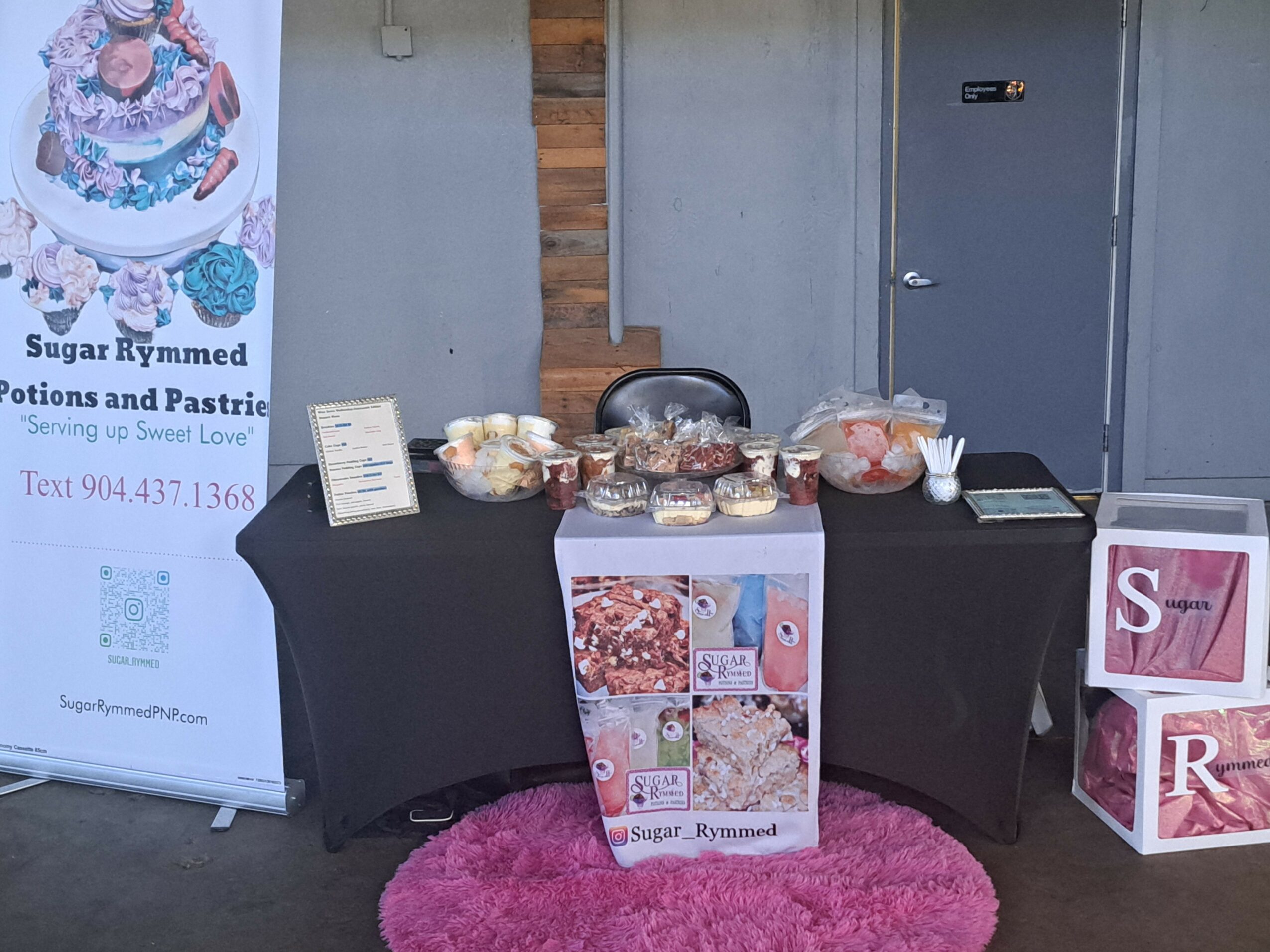 Table with baked goods and a sign, pink rug in front, poster on the left, and decorative blocks on the right.