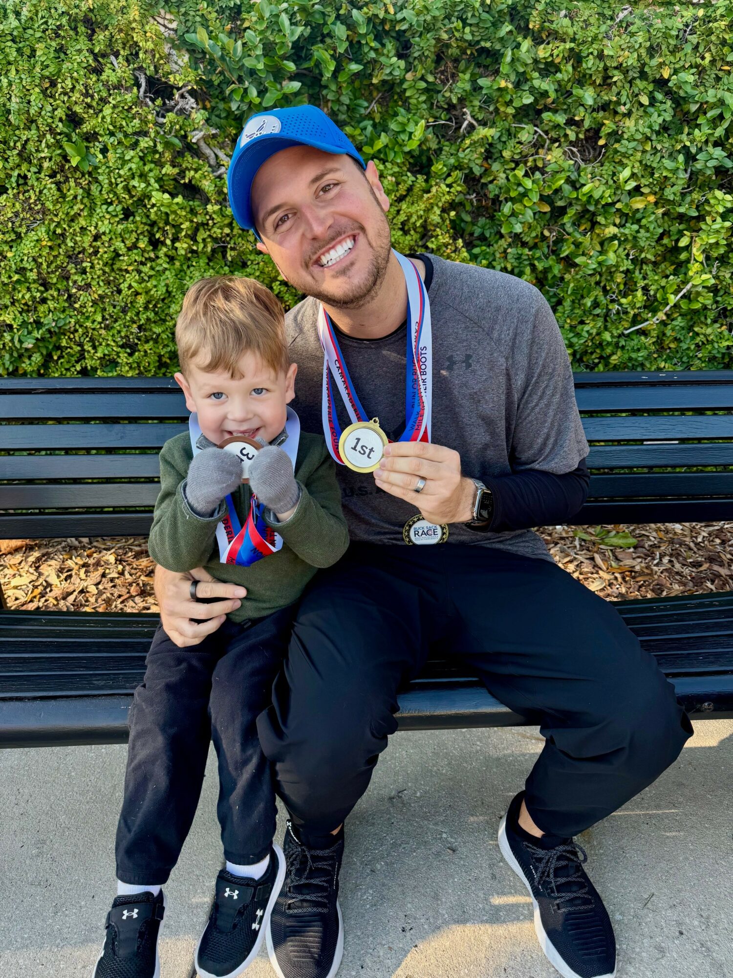 A smiling man and young boy sitting on a bench outdoors, both wearing medals and casual clothing, with green foliage behind them.