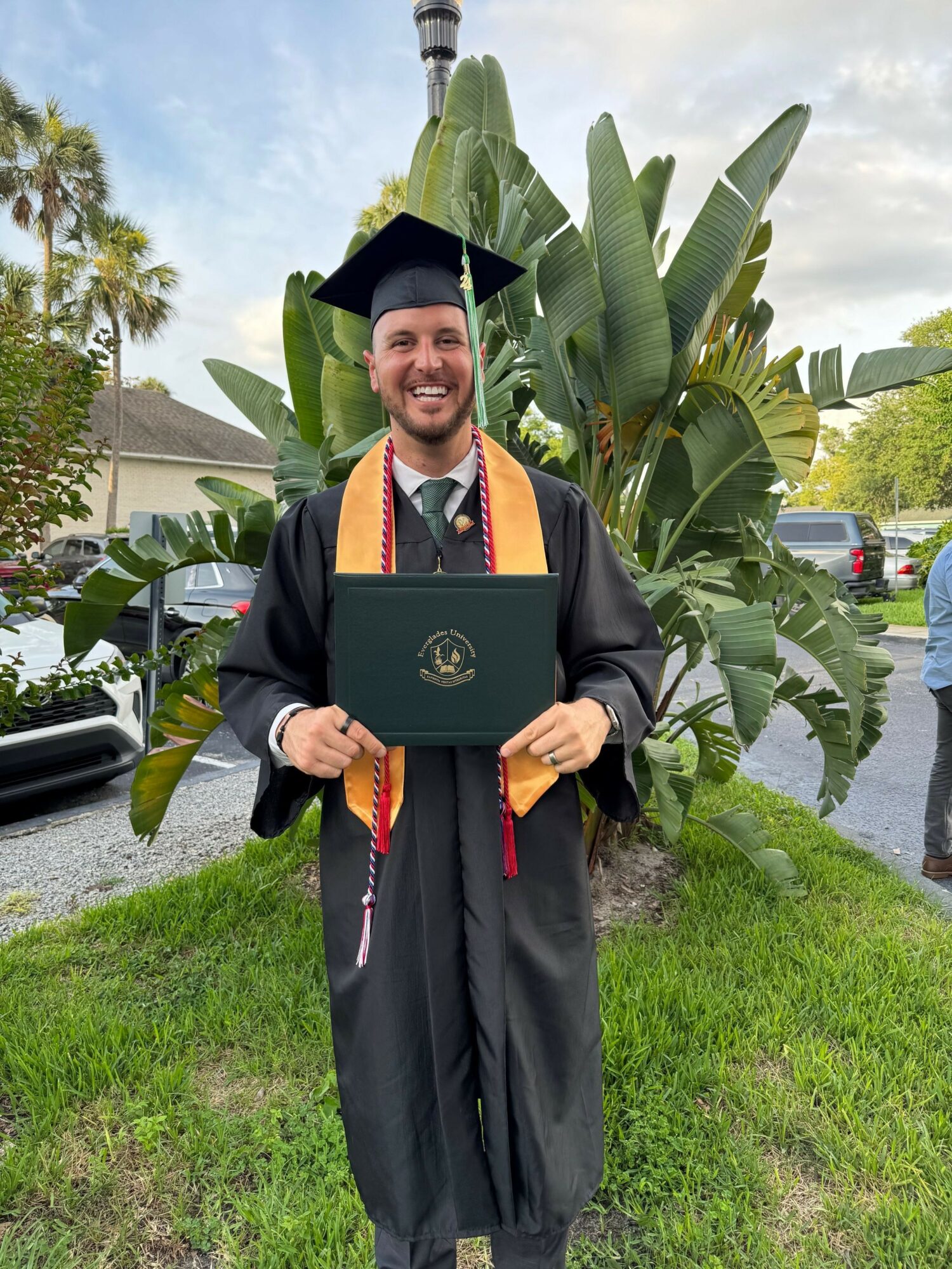 Graduate in cap and gown holding diploma outdoors with green plants and a street in background.