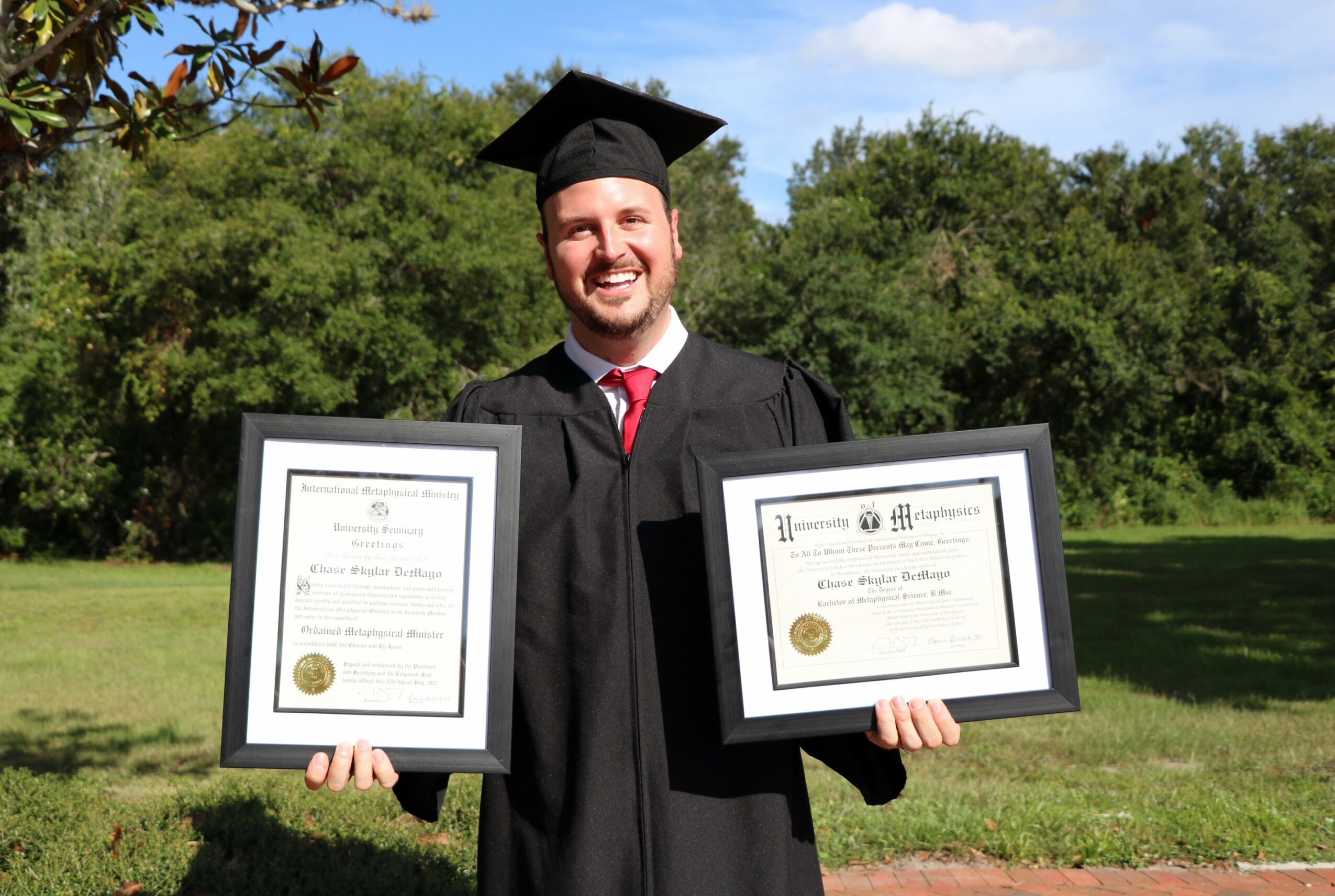 Graduate in cap and gown holding two framed certificates outdoors with trees and blue sky behind him.