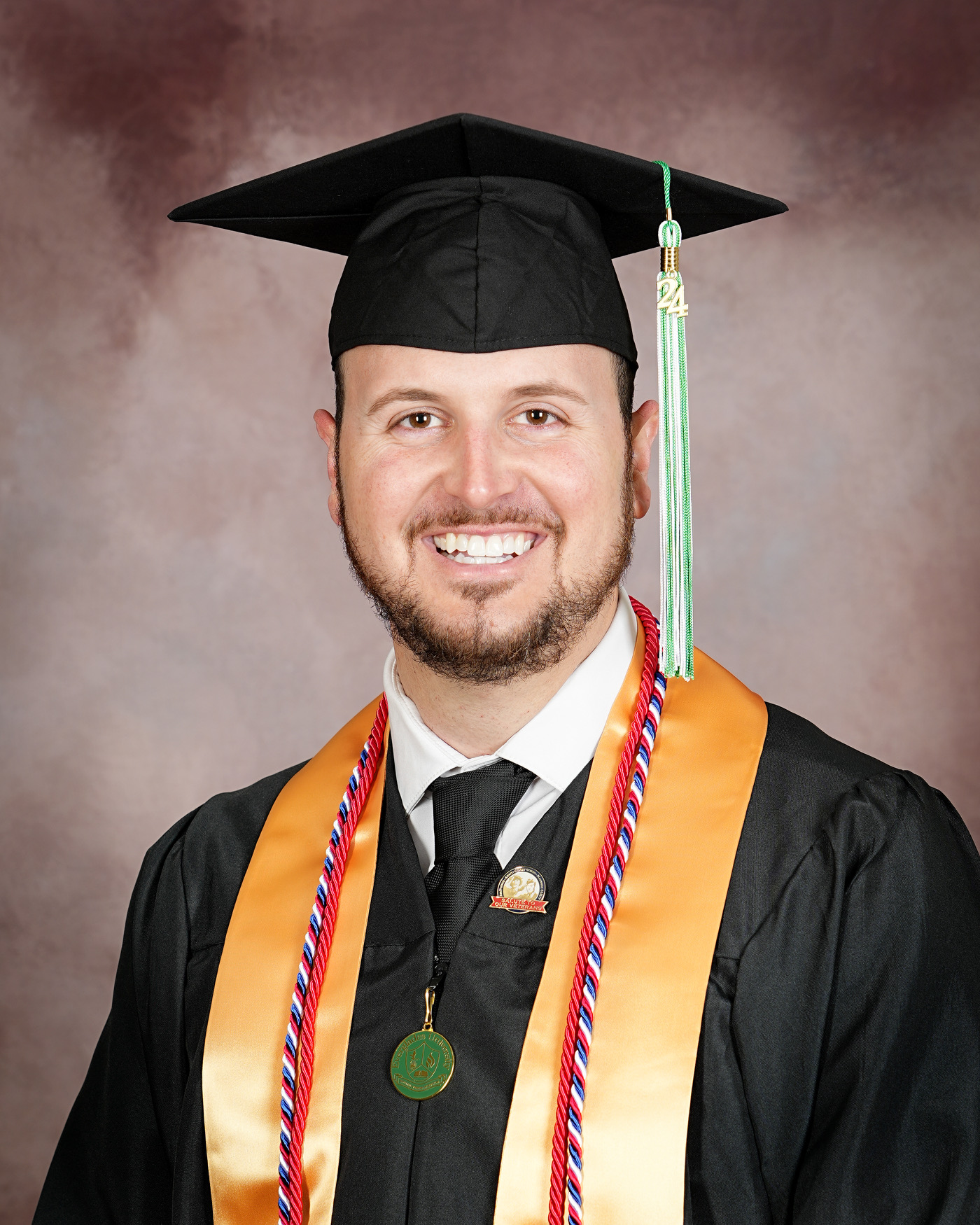 Man in graduation cap and gown smiling, wearing honor cords and medals, against a neutral background.