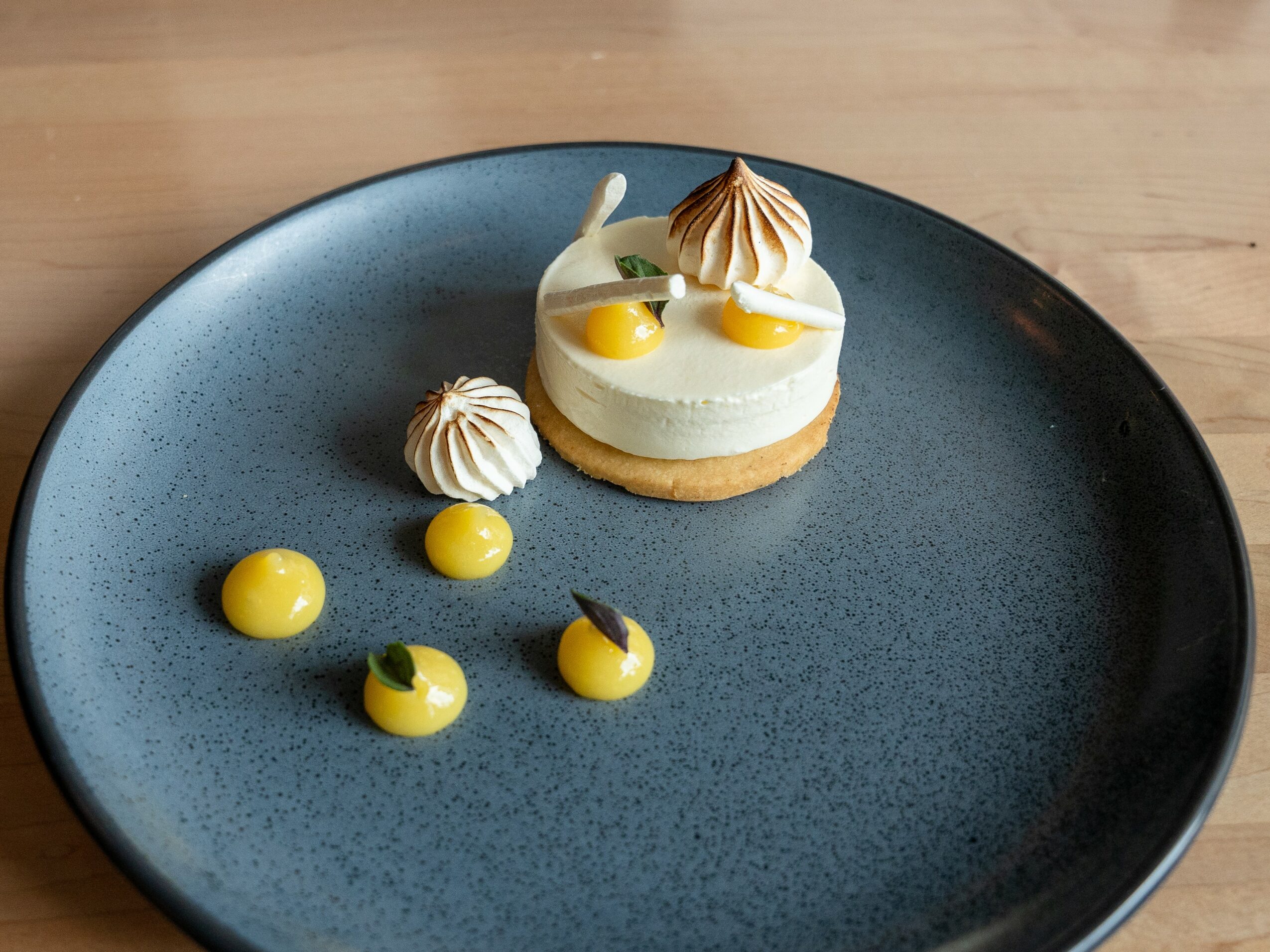 A dessert with cream, meringue, and yellow sauce on a dark blue plate, on a wooden surface.
