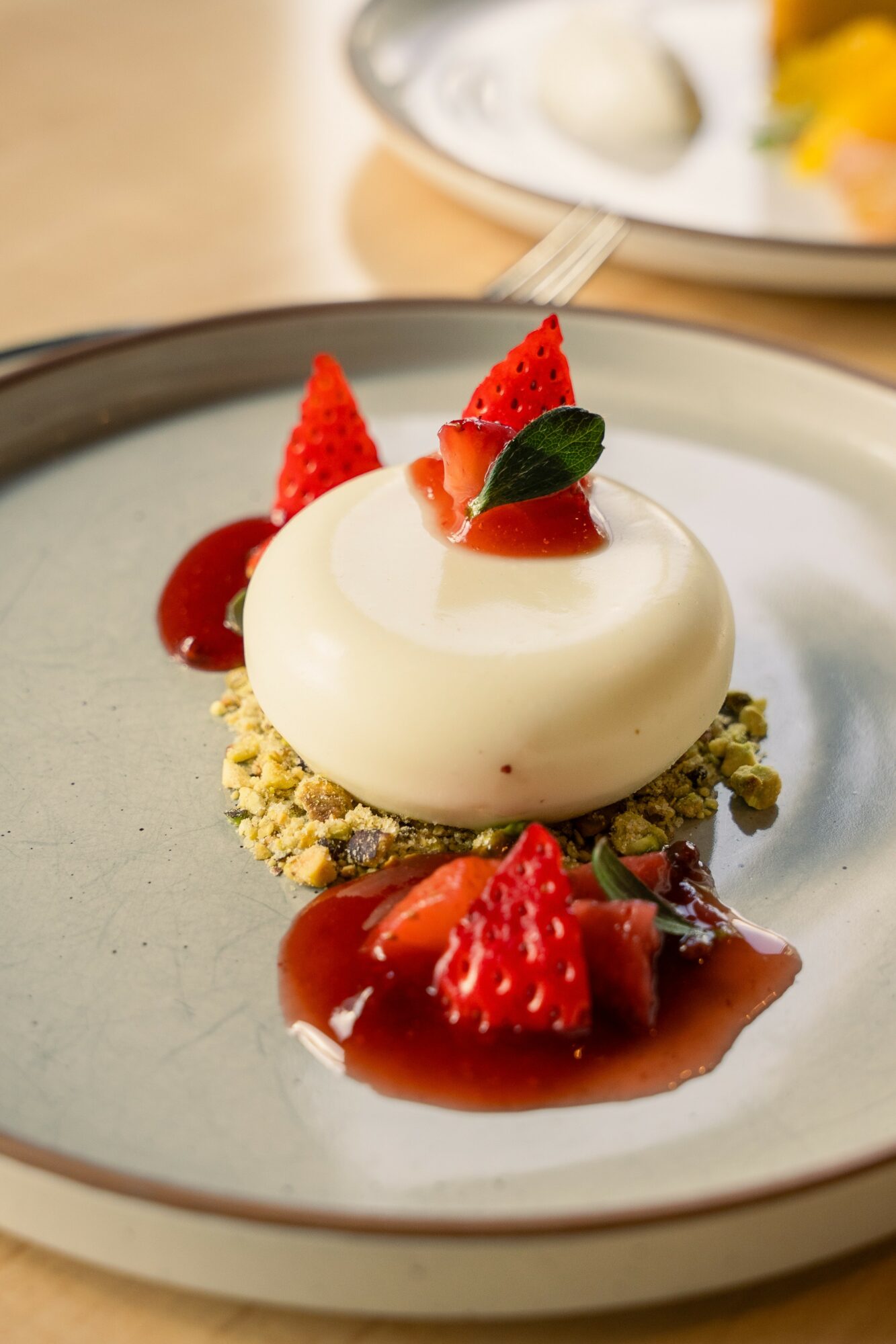 Dessert with white mousse, strawberries, and green leaf on a gray plate, with a blurred background.