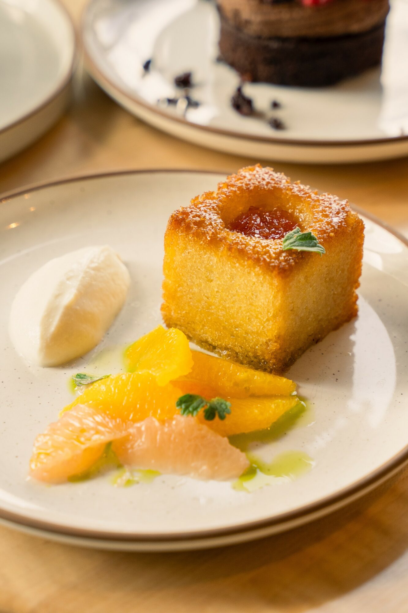 Square yellow cake with a fruit topping, served with citrus slices and a dollop of cream on a white plate.