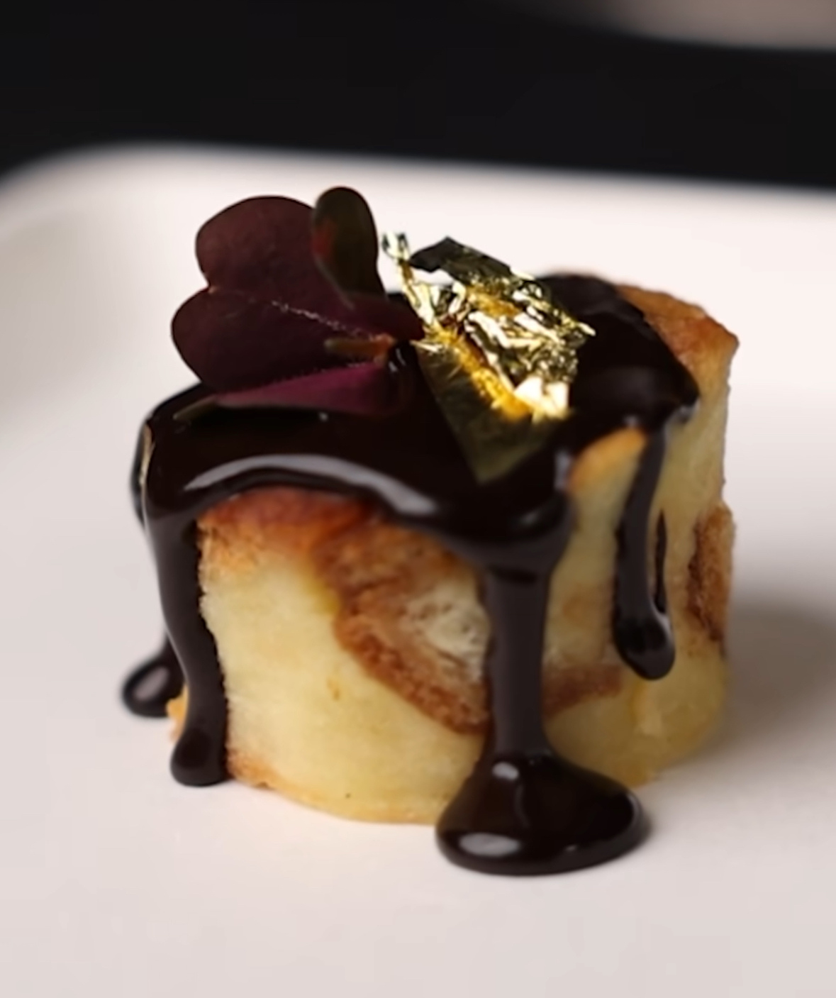 Cake with dark chocolate glaze, garnished with edible gold leaf and purple leaves, on a white plate.