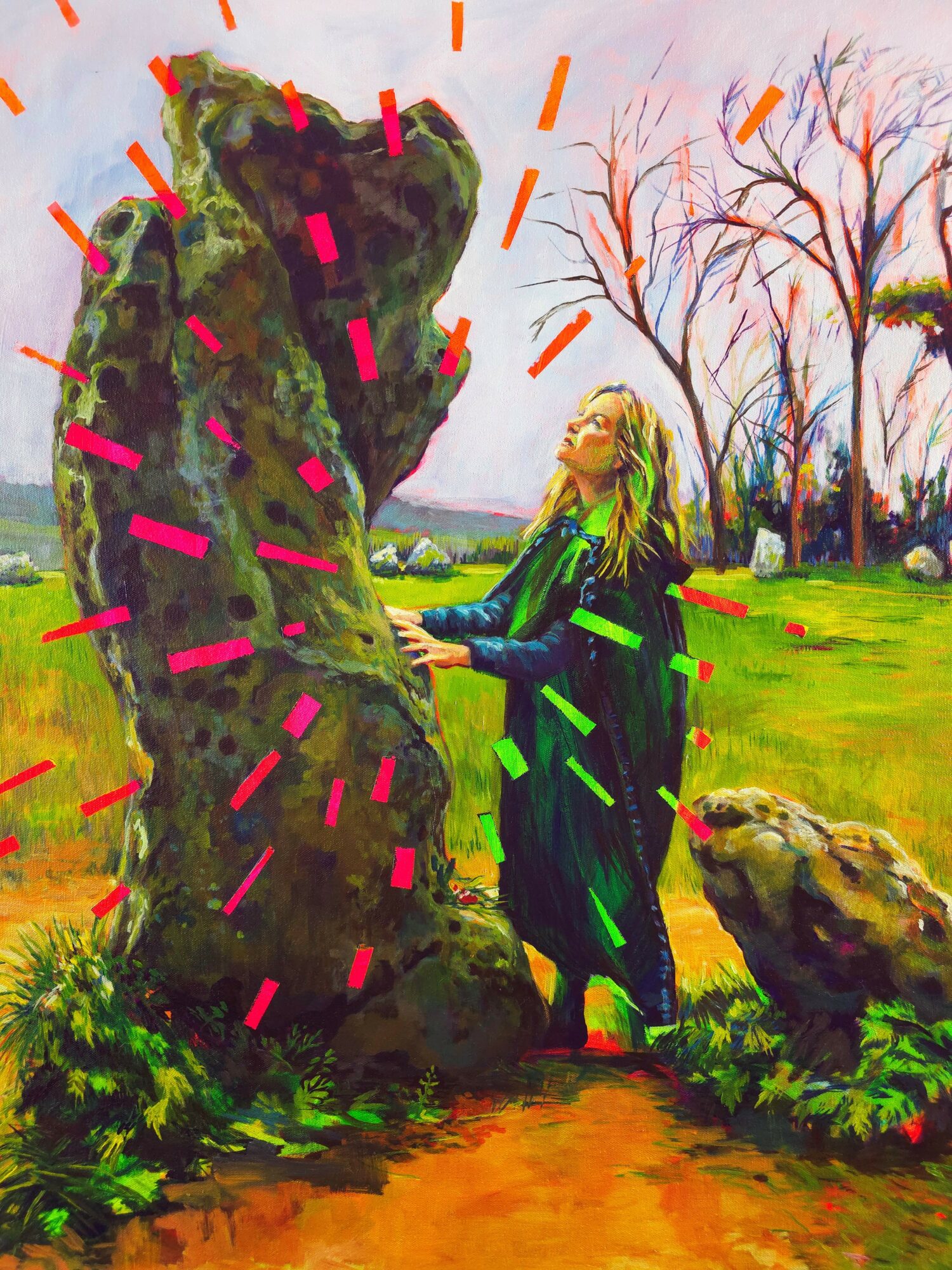 Girl with long blonde hair in a green dress touches a large moss-covered rock in a park with trees and grass.