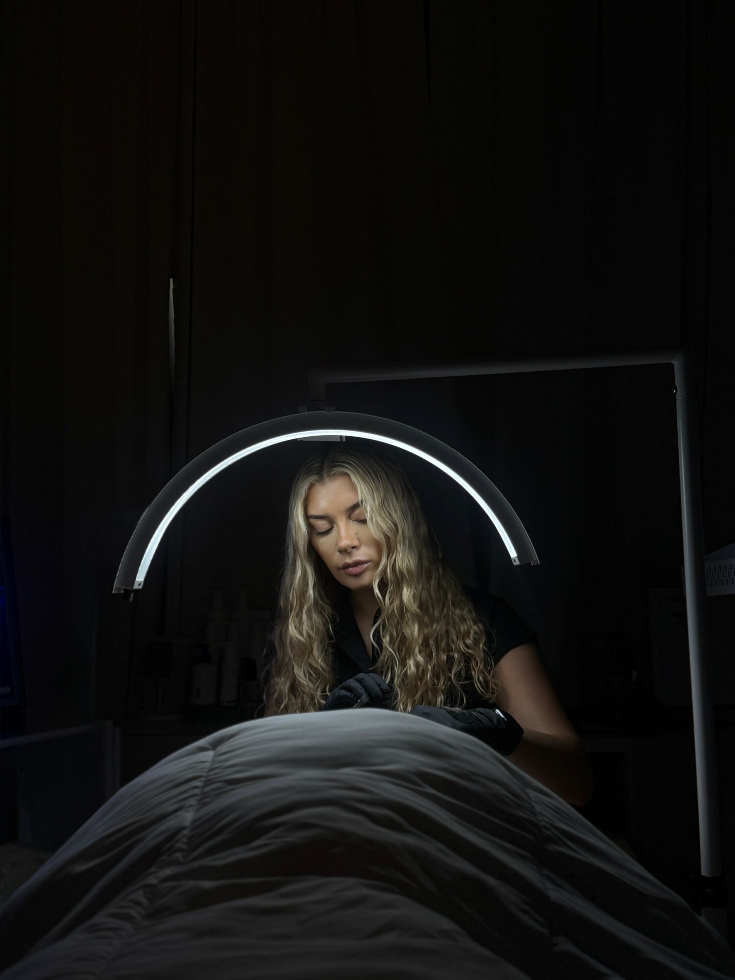 Young woman with long wavy hair looks down, illuminated by a circular light, in a dark room with a bed in foreground.