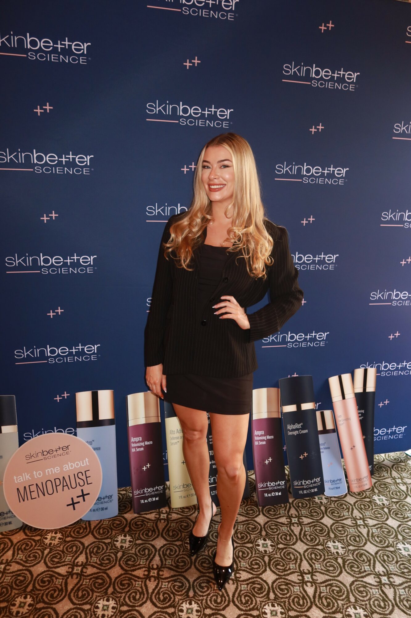 Woman with long blonde hair in black dress standing on patterned carpet in front of a blue backdrop with skincare brand logos, smiling.