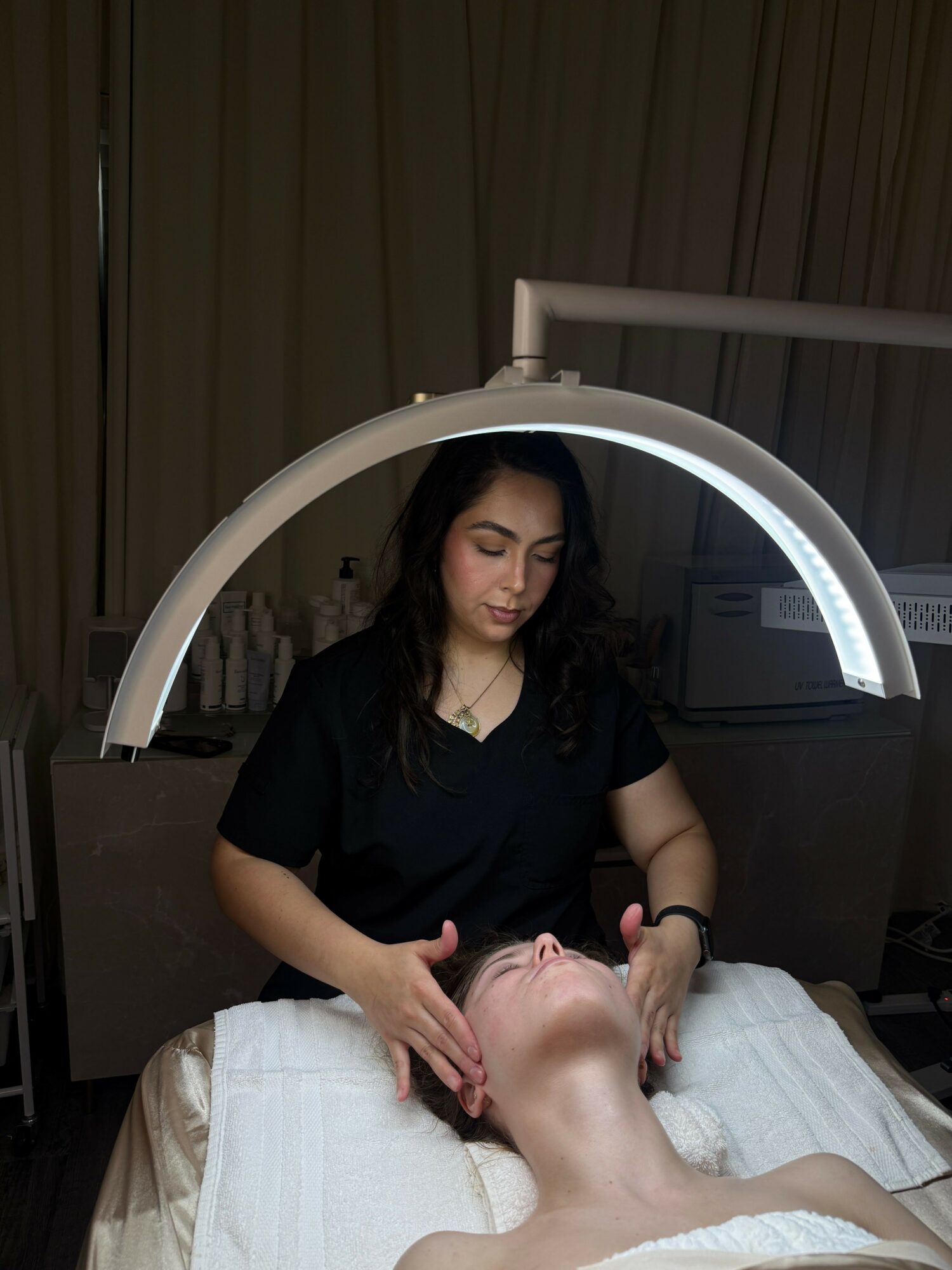 A woman receiving a facial treatment under a circular light, lying on a bed with a towel, in a spa or clinic setting.