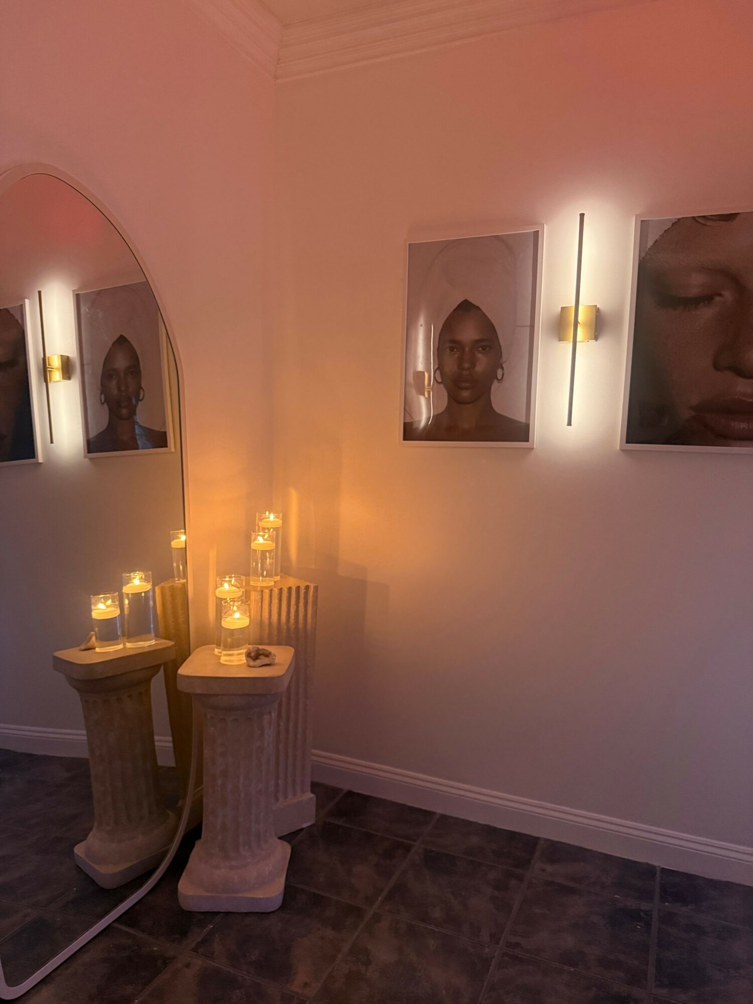 Upright view of a room corner with framed photos, a lit candle, and a decorative pedestal with candles and a sculpture.