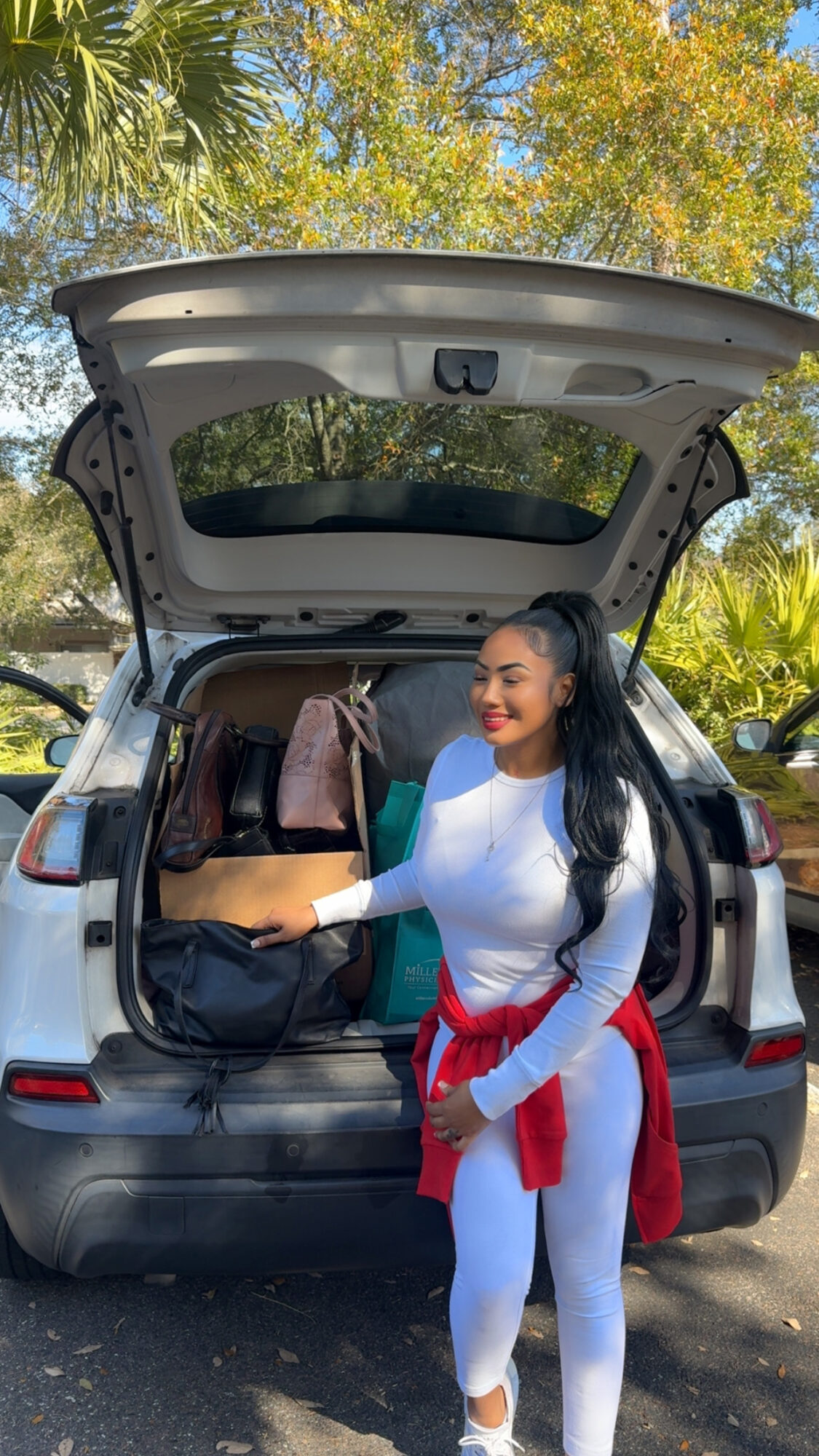 Woman standing next to an open car trunk outdoors with trees and sunlight, carrying a bag and wearing white athletic clothing.