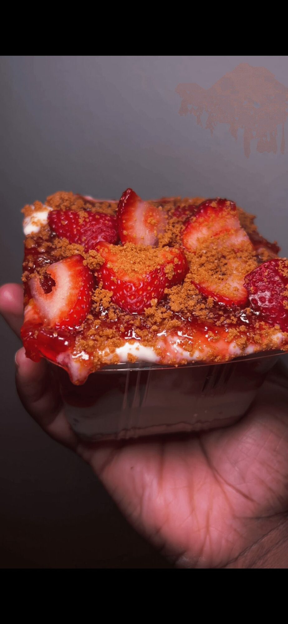 Hand holding a slice of strawberry-topped cake with colorful sprinkles against a plain background.