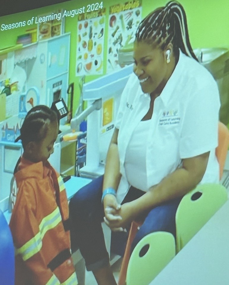 A woman in a white shirt interacts with a child in an orange jacket in a colorful classroom setting.