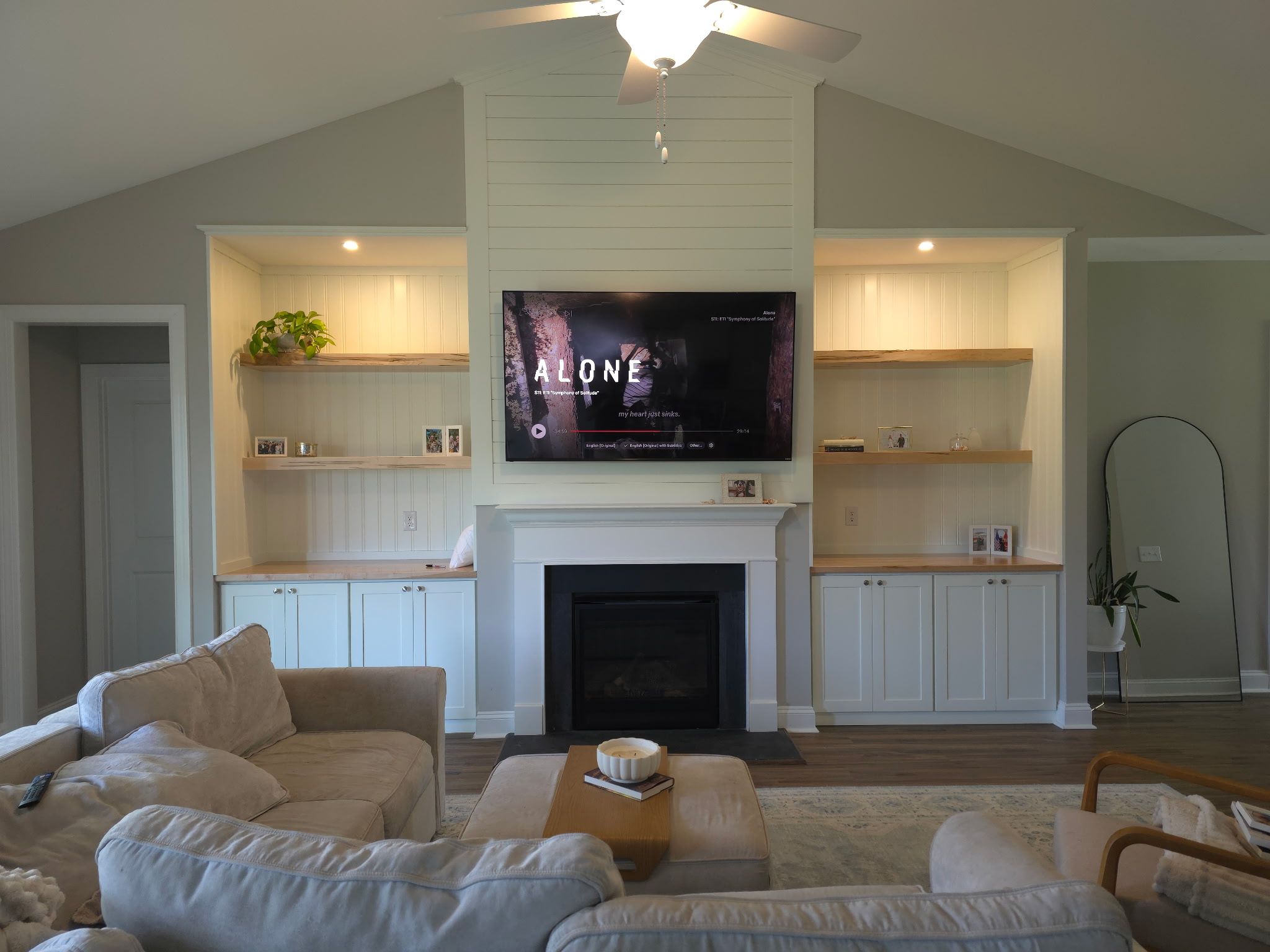 Living room with built-in shelves, a fireplace, a wall-mounted TV, and a beige sofa, with a mirror and plant nearby.