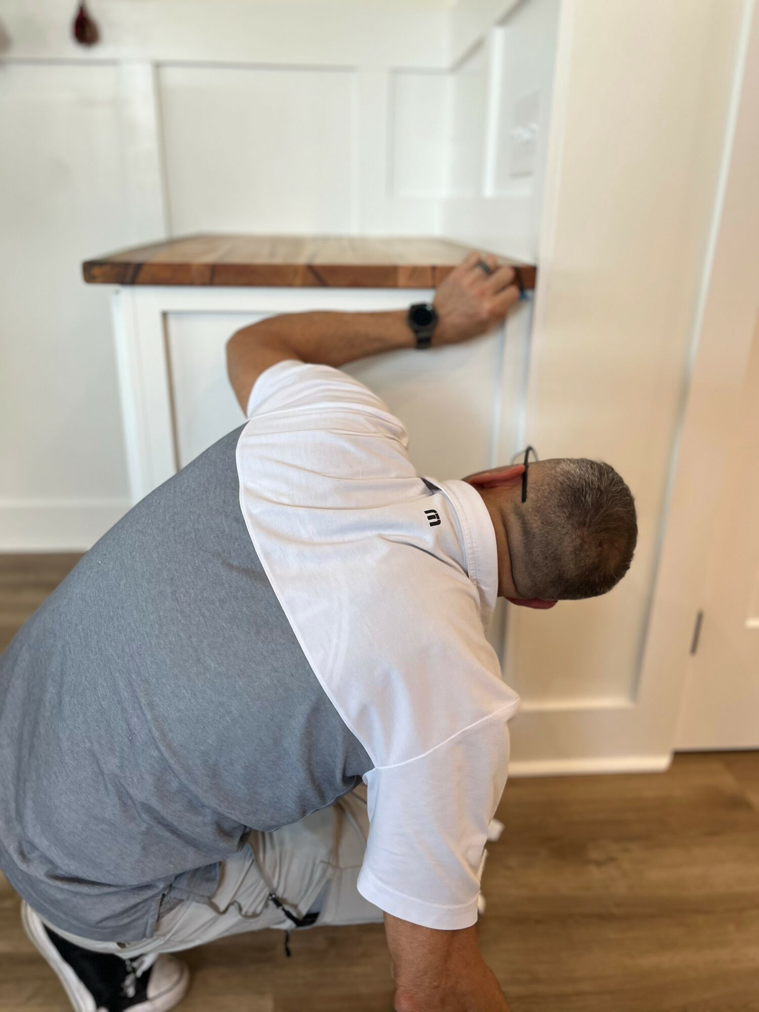 Man leaning over a kitchen counter, reaching with his right hand, wearing a gray vest and white shirt, bald head.