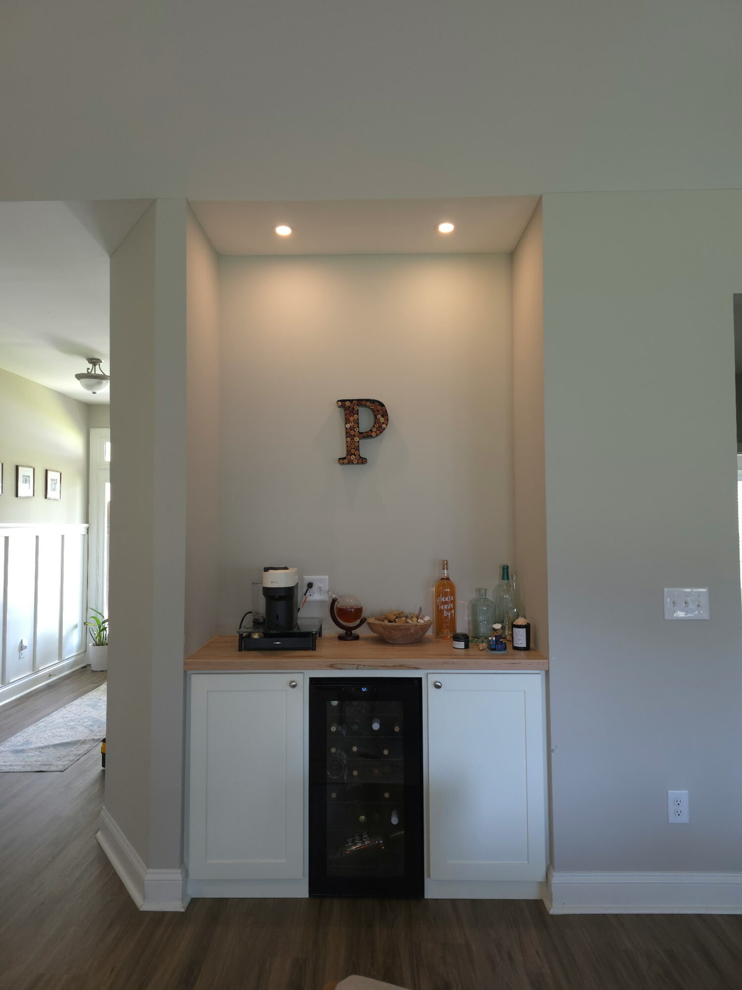 Built-in bar area with a small wine fridge, coffee maker, and bottles, illuminated by two ceiling lights, against a white wall.